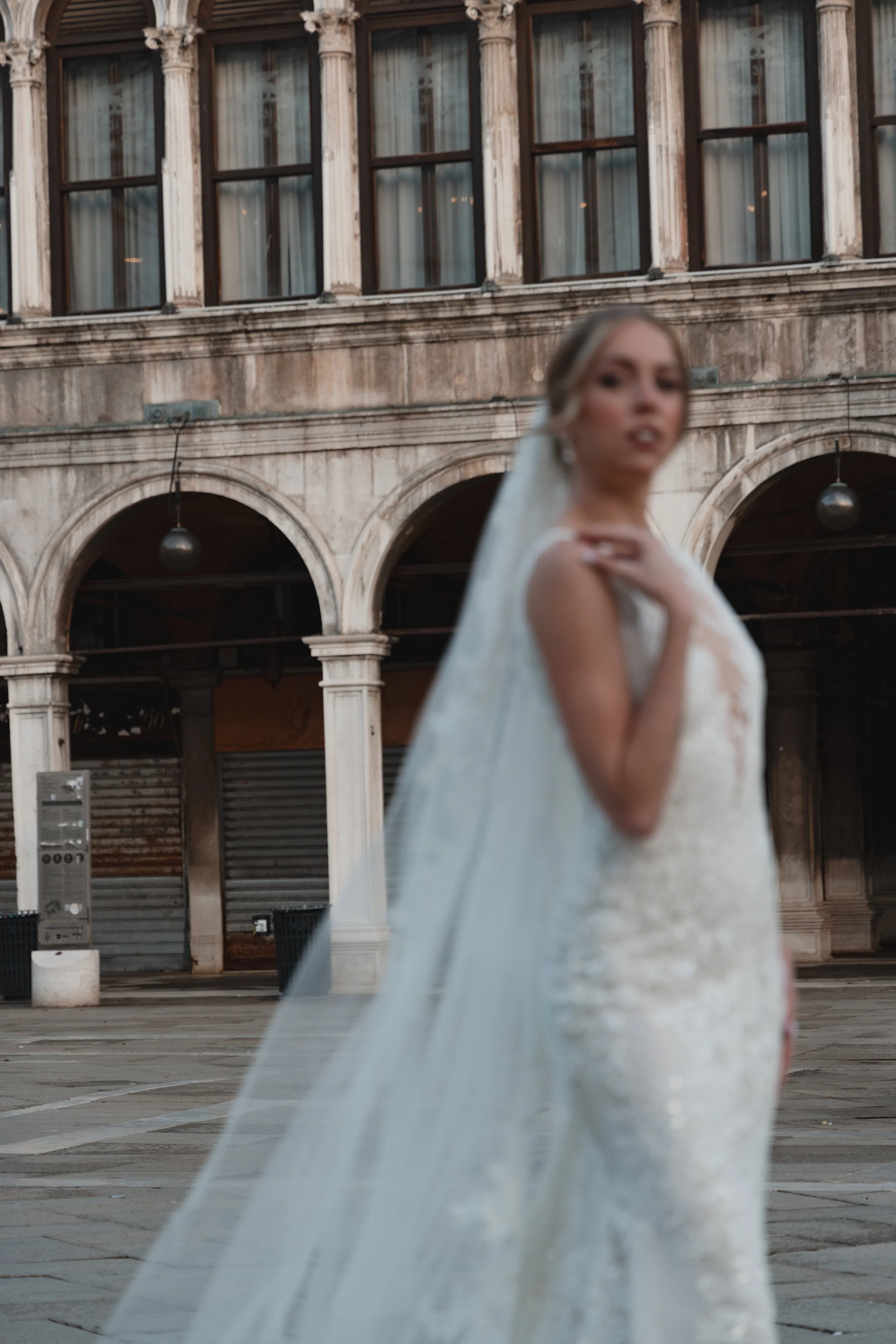 Blurry image of a woman in a wedding dress standing outdoors in front of an old brick building with arched windows and classical columns.