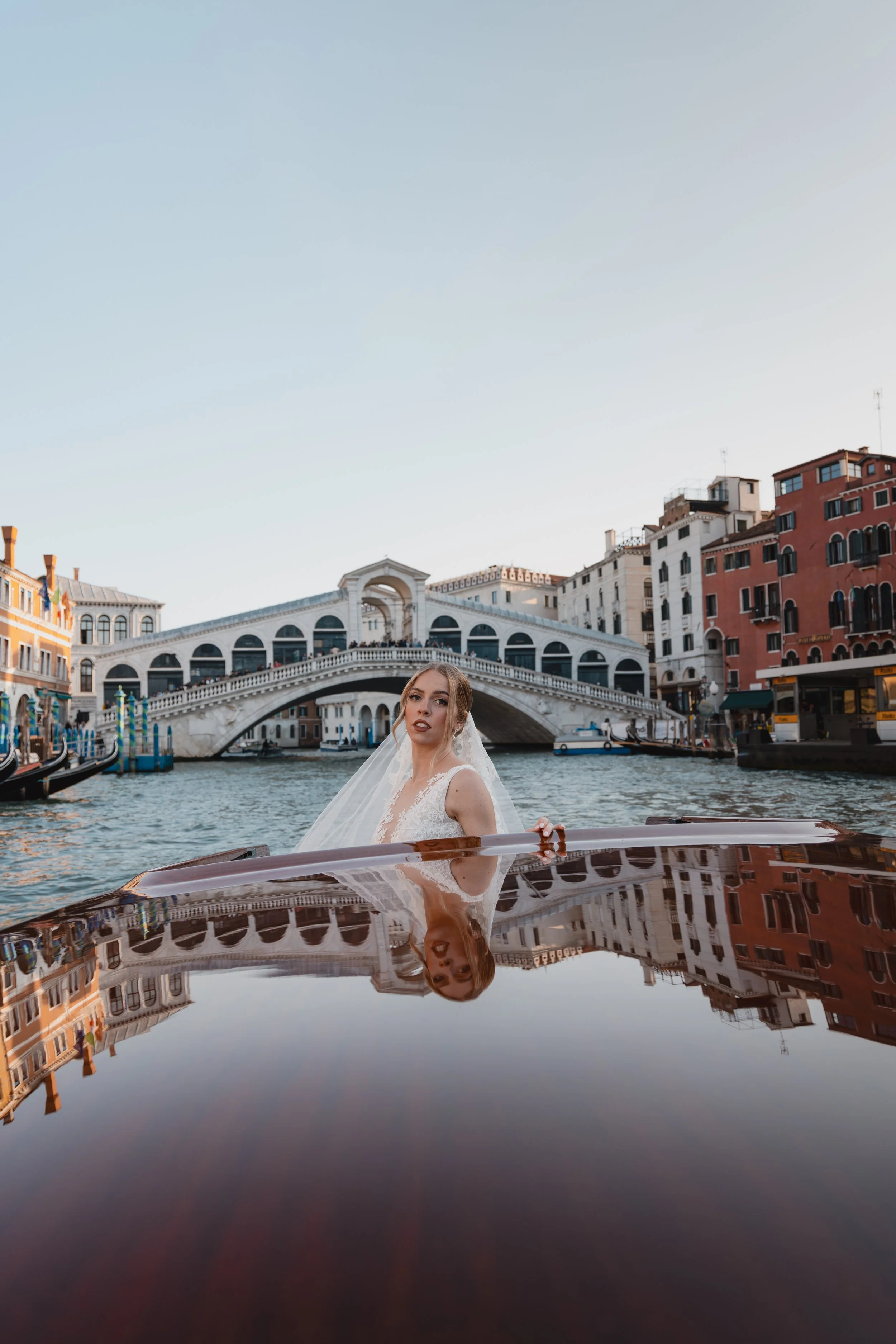 A bride in a white wedding dress and veil is on a boat in a canal, with historic buildings and a bridge in Venice, Italy, in the background. The water's reflection creates a mirror image of her and the scene.