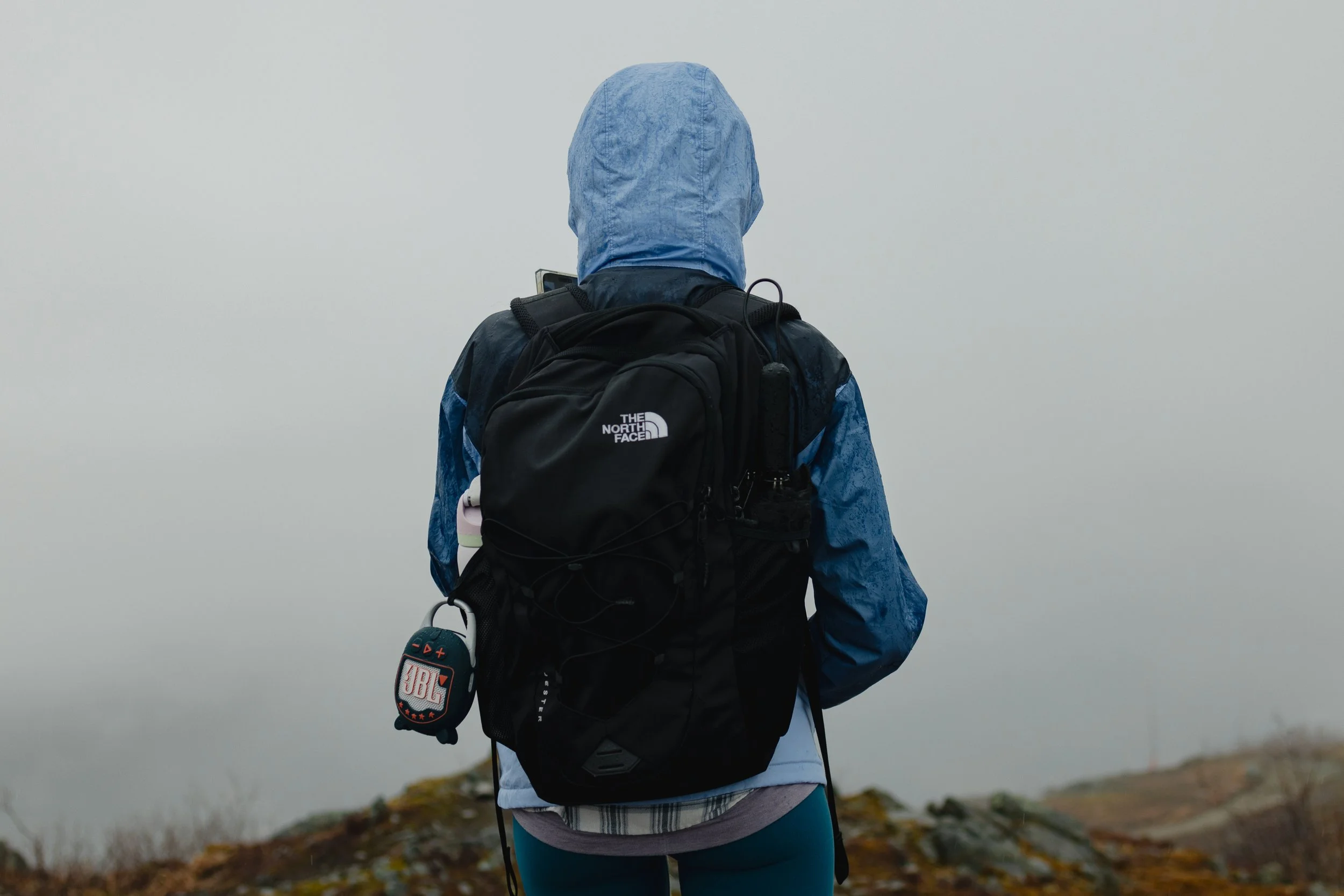 A person dressed in a blue hooded jacket, carrying a black backpack, is hiking outdoors near a body of water on a cloudy day.