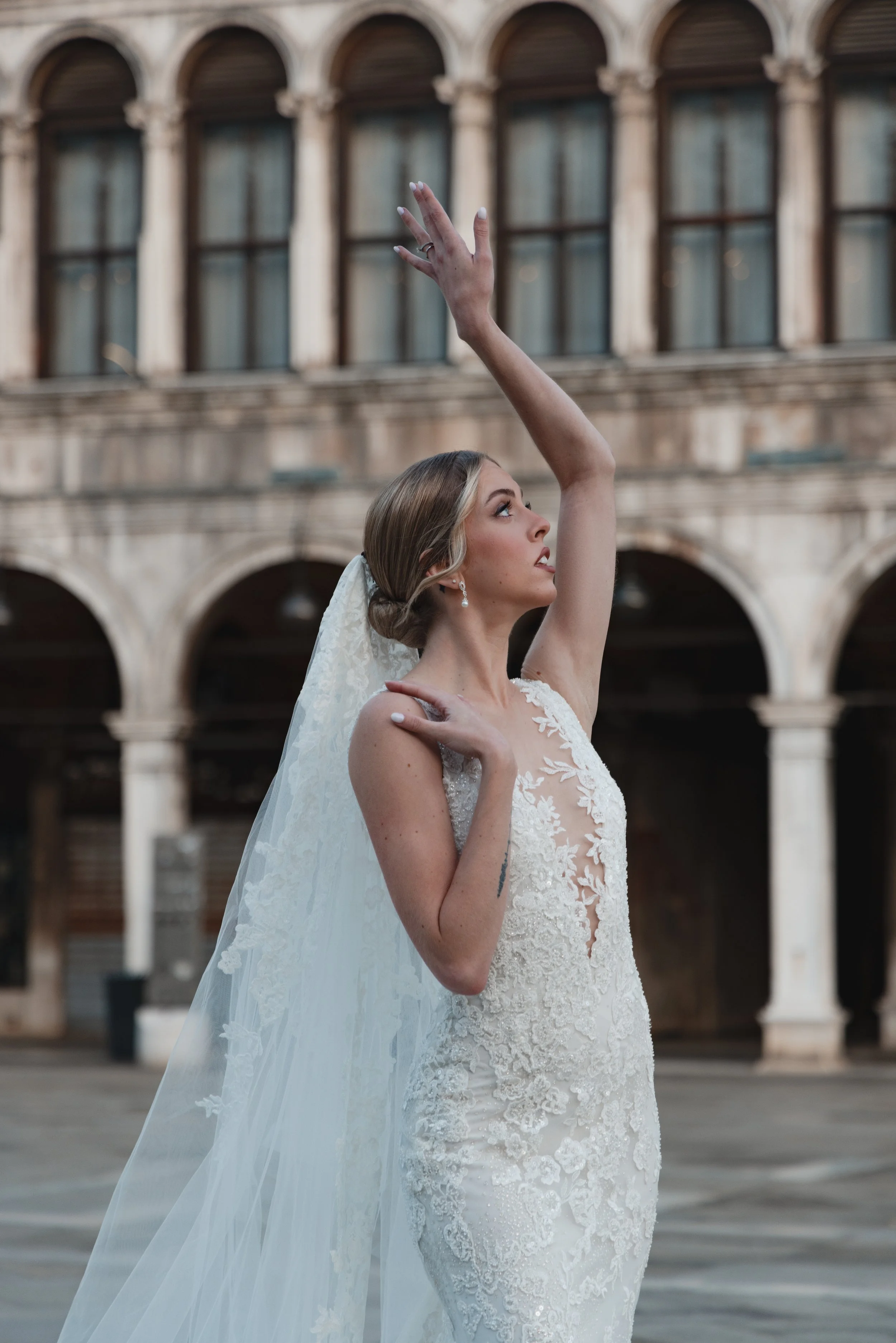 A bride in a white lace wedding dress and veil dances outside in front of an old stone building with arched windows.