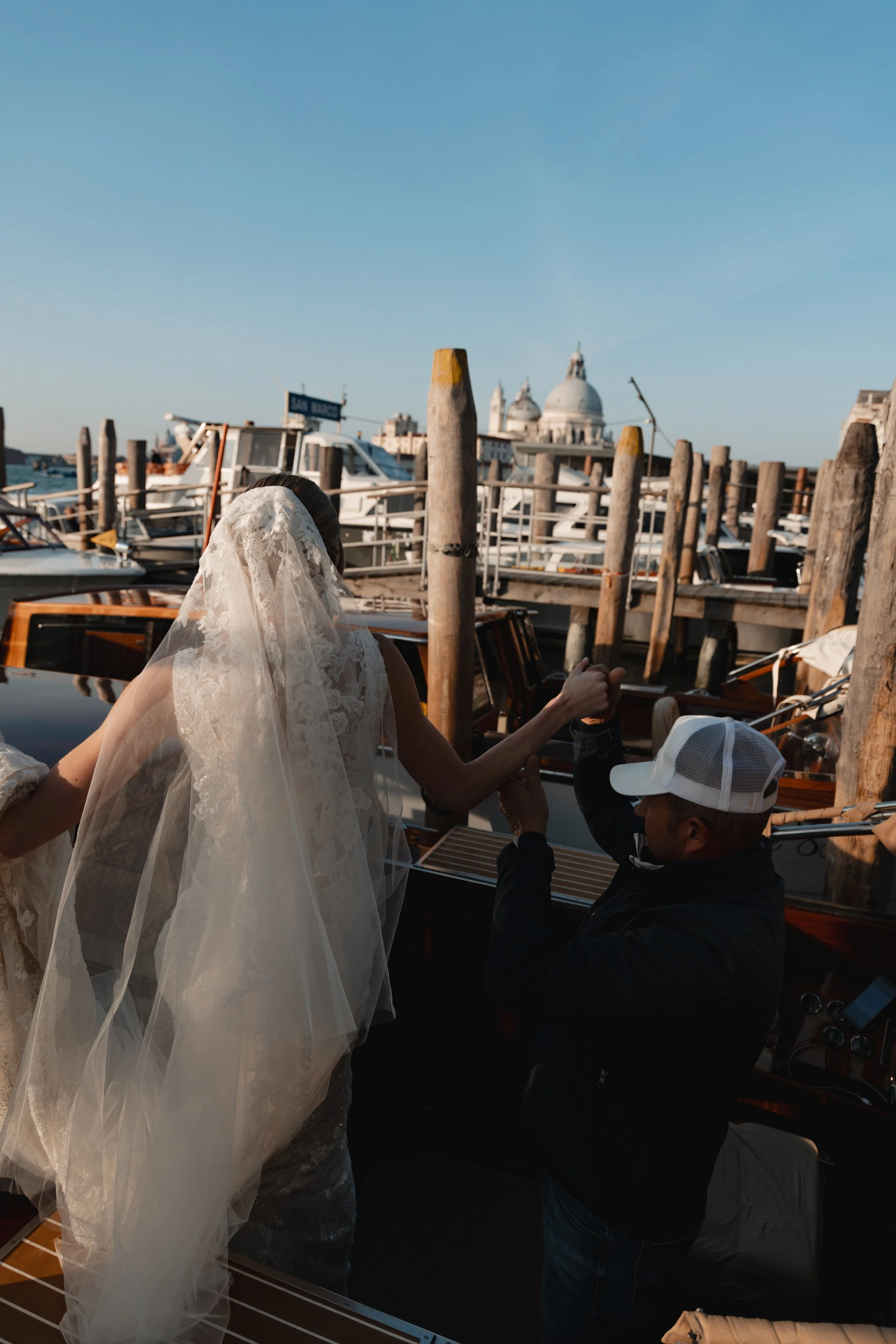 A woman in a white wedding dress and veil is on a boat dock, holding hands with a man wearing a baseball cap and black jacket. In the background, there are boats and a cityscape with a domed building.