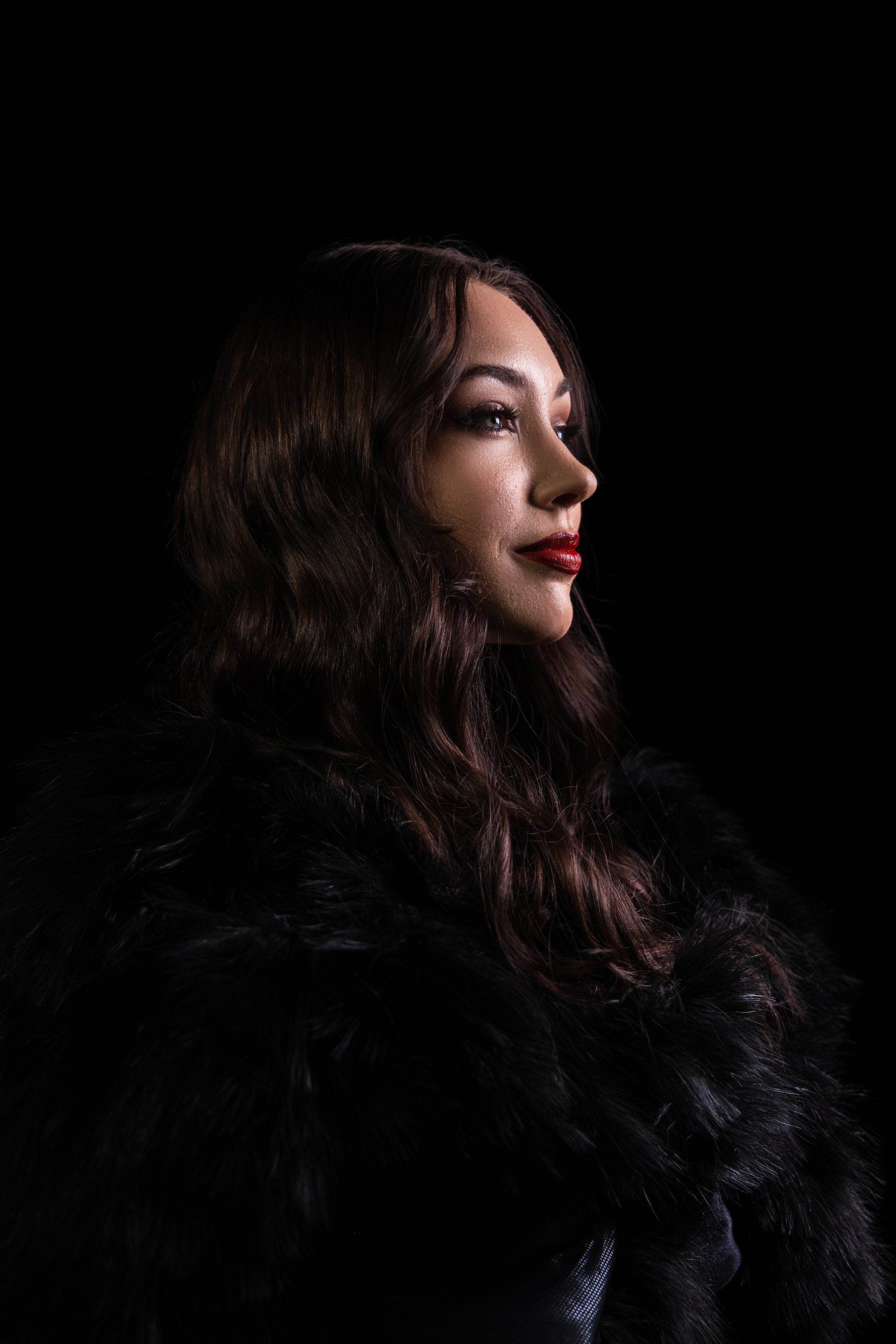 A woman with wavy brown hair, red lipstick, and makeup, wearing a black fur coat, with a dark background.