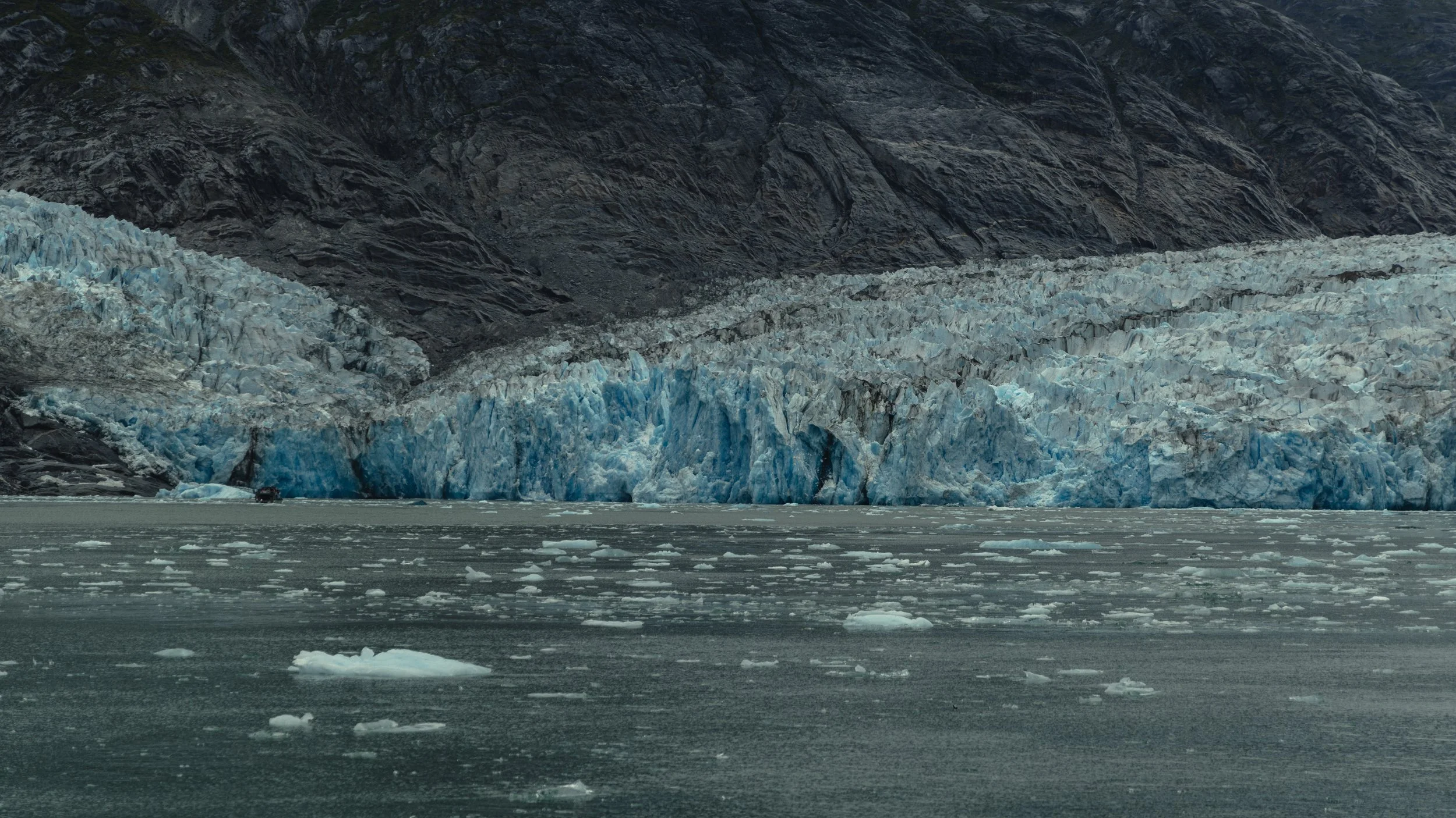A large glacier with blue and white ice extending from a mountainous landscape into a body of water with floating ice chunks.