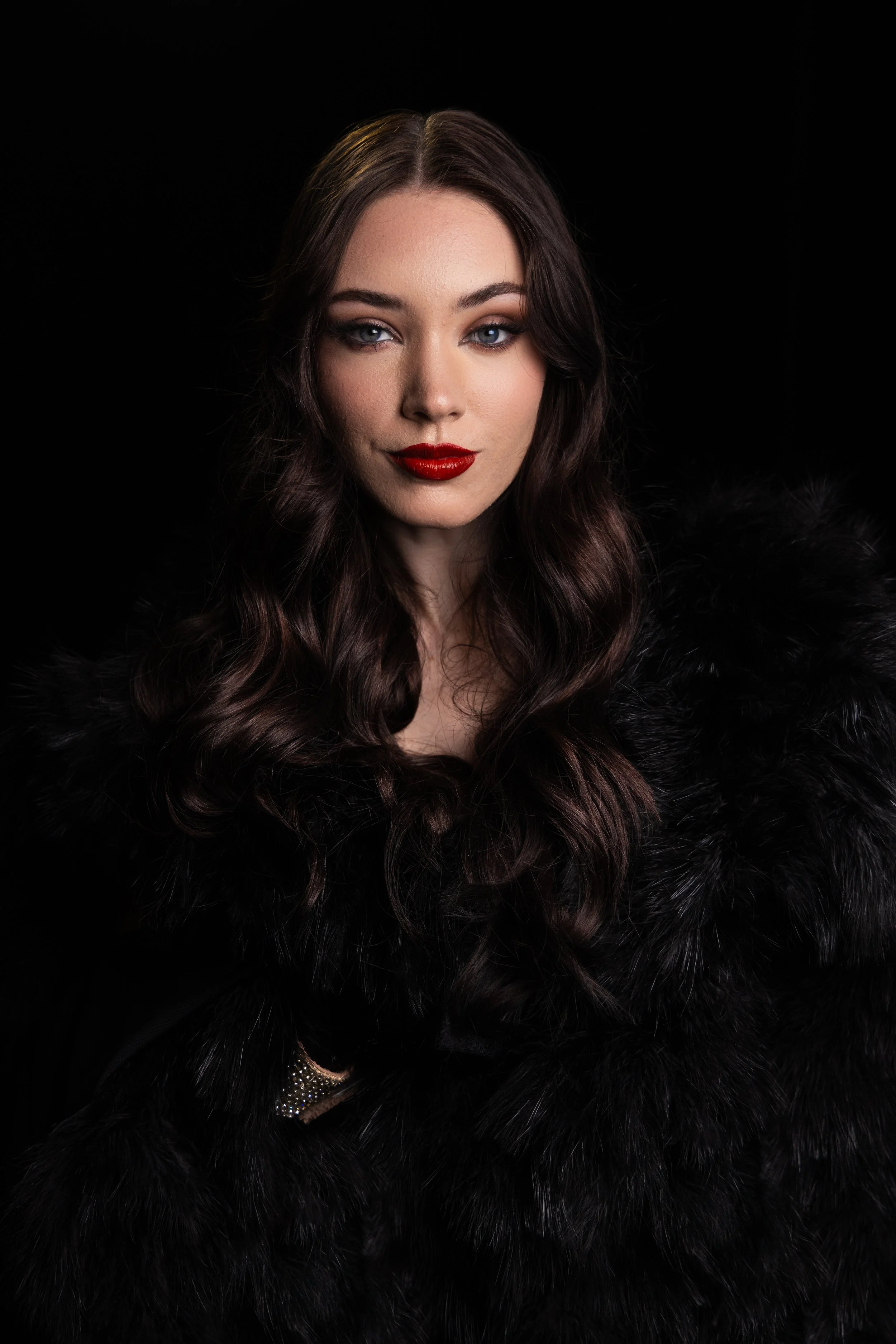 A woman with long, wavy dark hair, blue eyes, wearing red lipstick, a black fur coat, and posing against a black background.