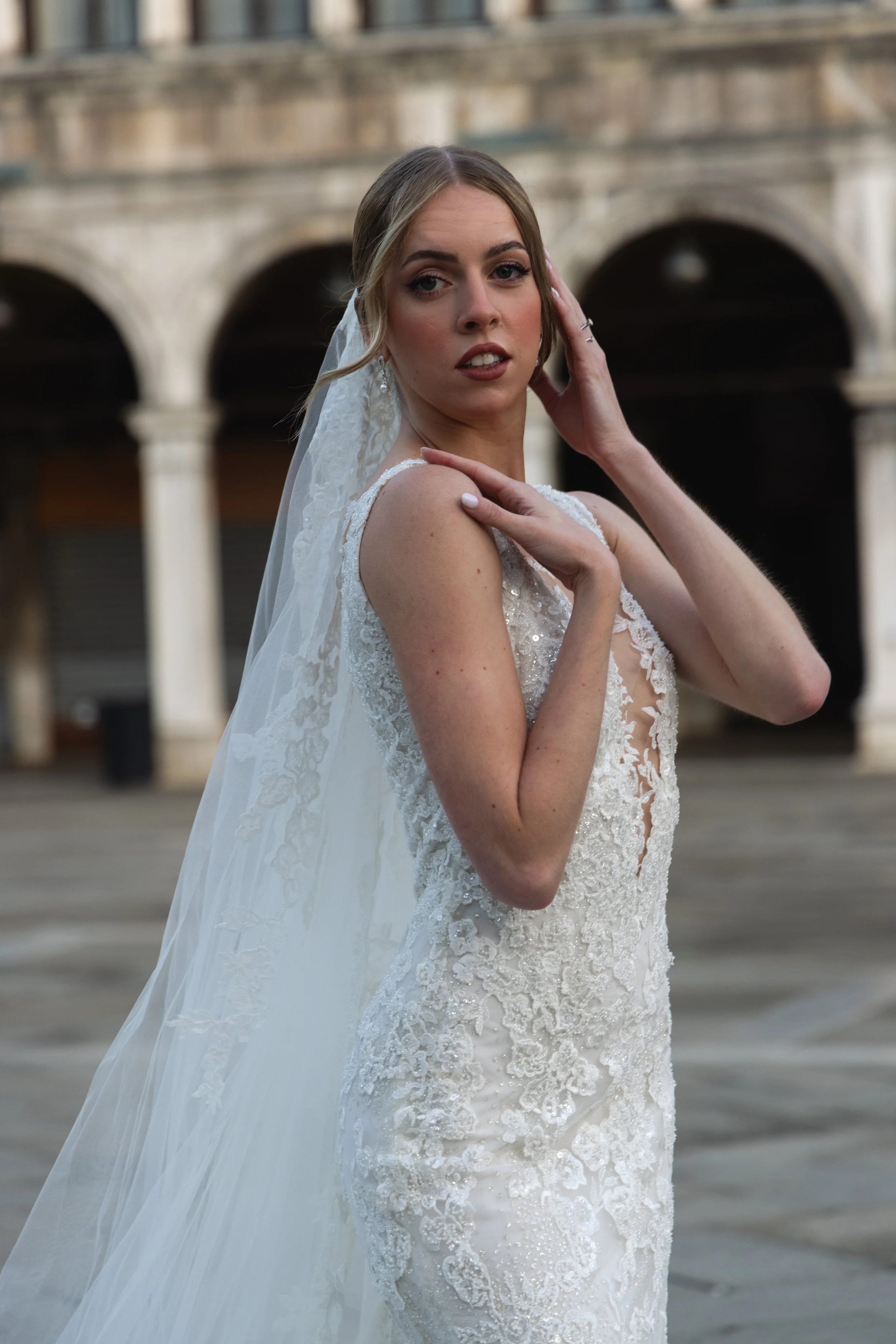A young woman in a wedding dress with lace details, wearing a veil, stands outside with an urban background featuring stone arches, touching her face and looking into the camera.