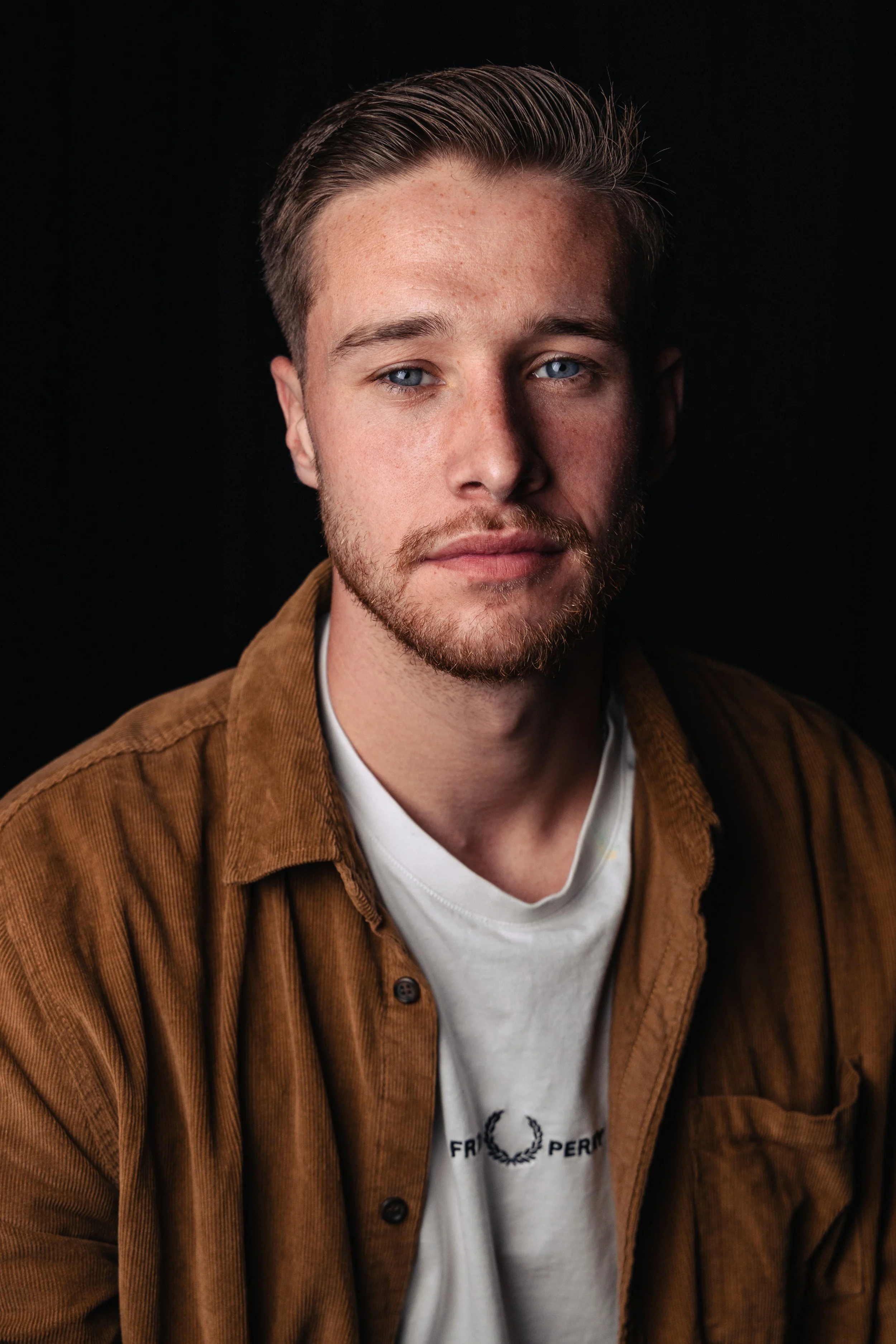 Close-up portrait of a young man with blue eyes, light skin, short wavy hair, and a beard, wearing a brown corduroy jacket over a white T-shirt, against a black background.