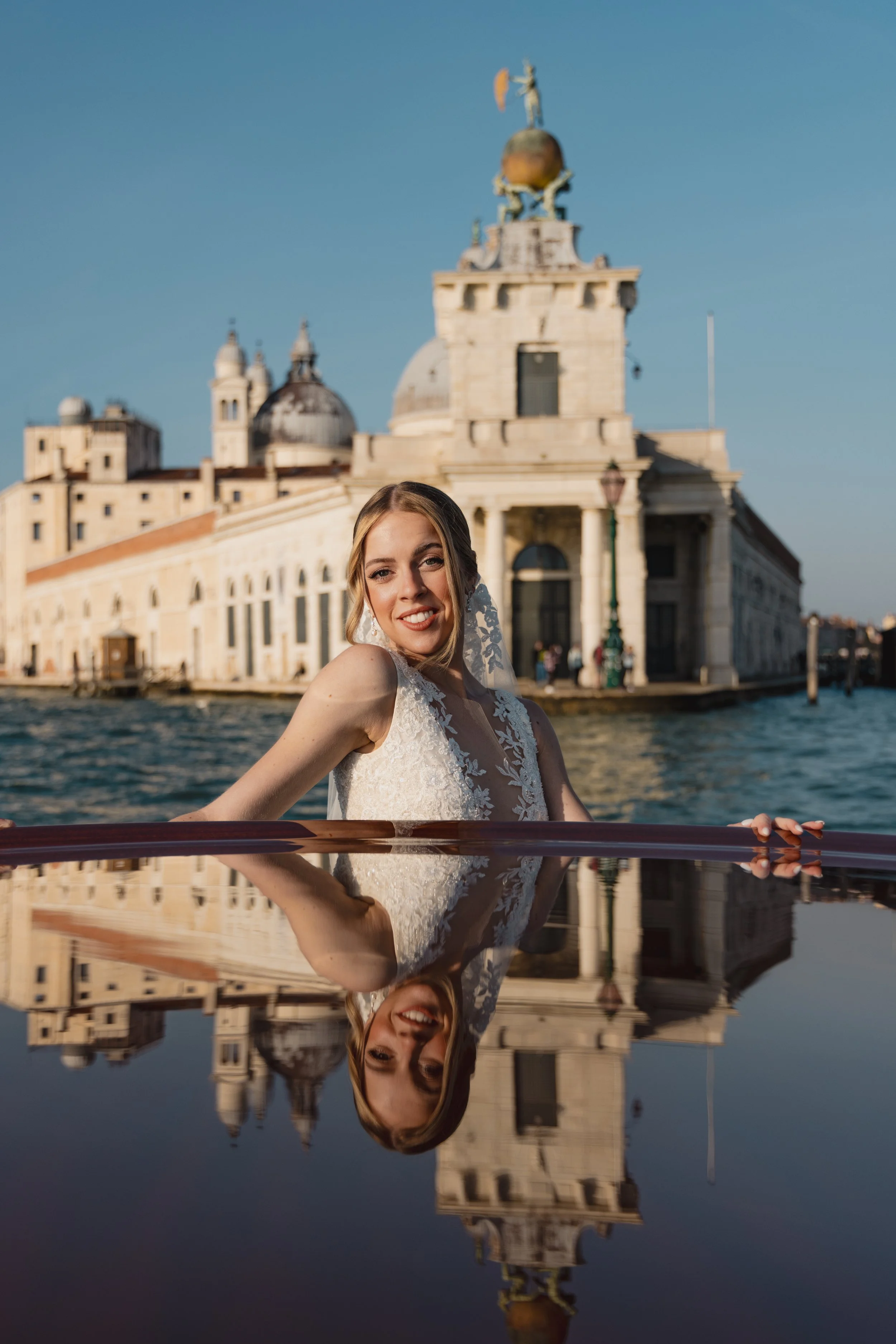 A woman in wedding attire smiling at the camera, with a historic building and water in the background, and her reflection visible in the water.