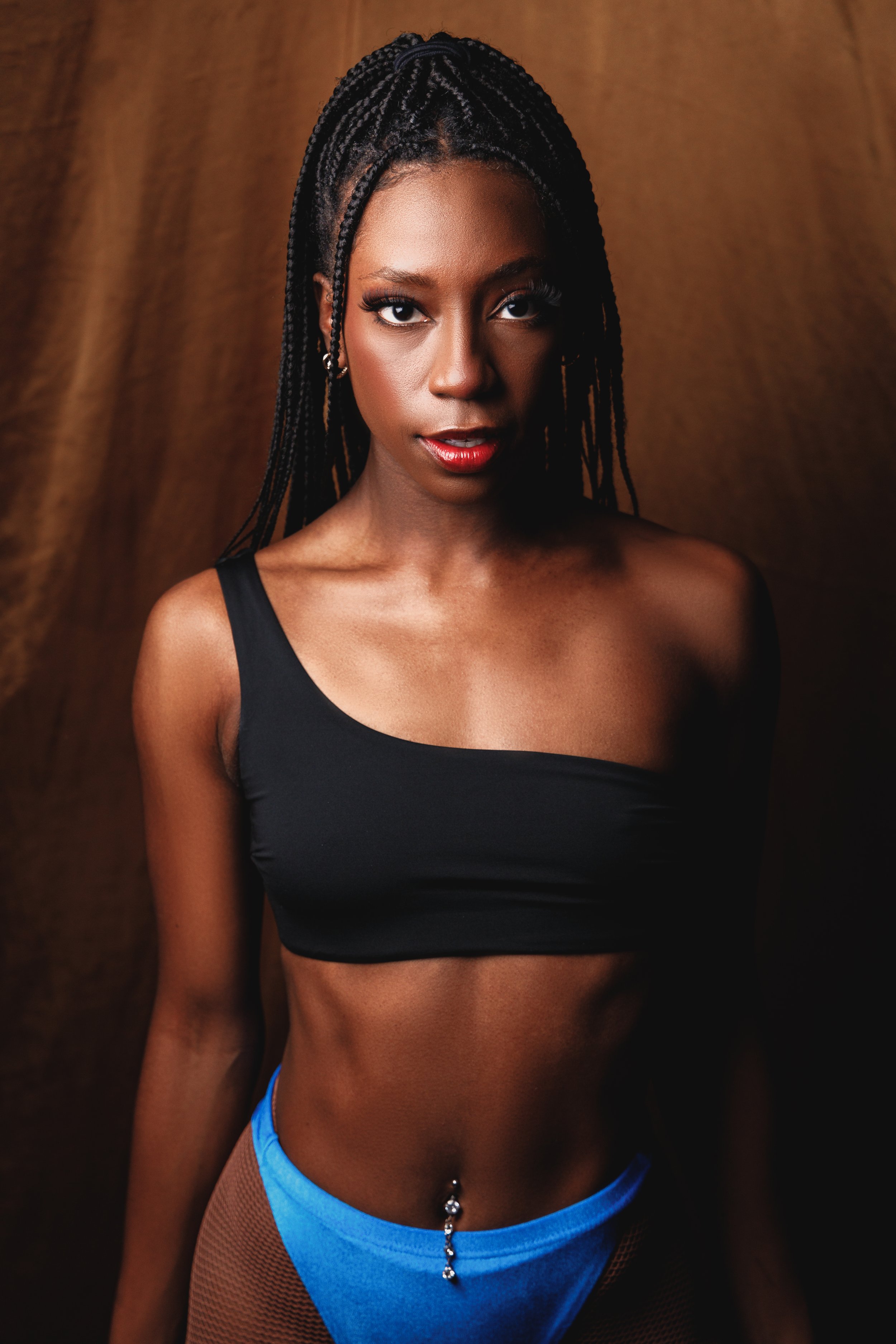 Close-up of a woman with braids, wearing a black sports bra and blue athletic underwear, with a brown background.