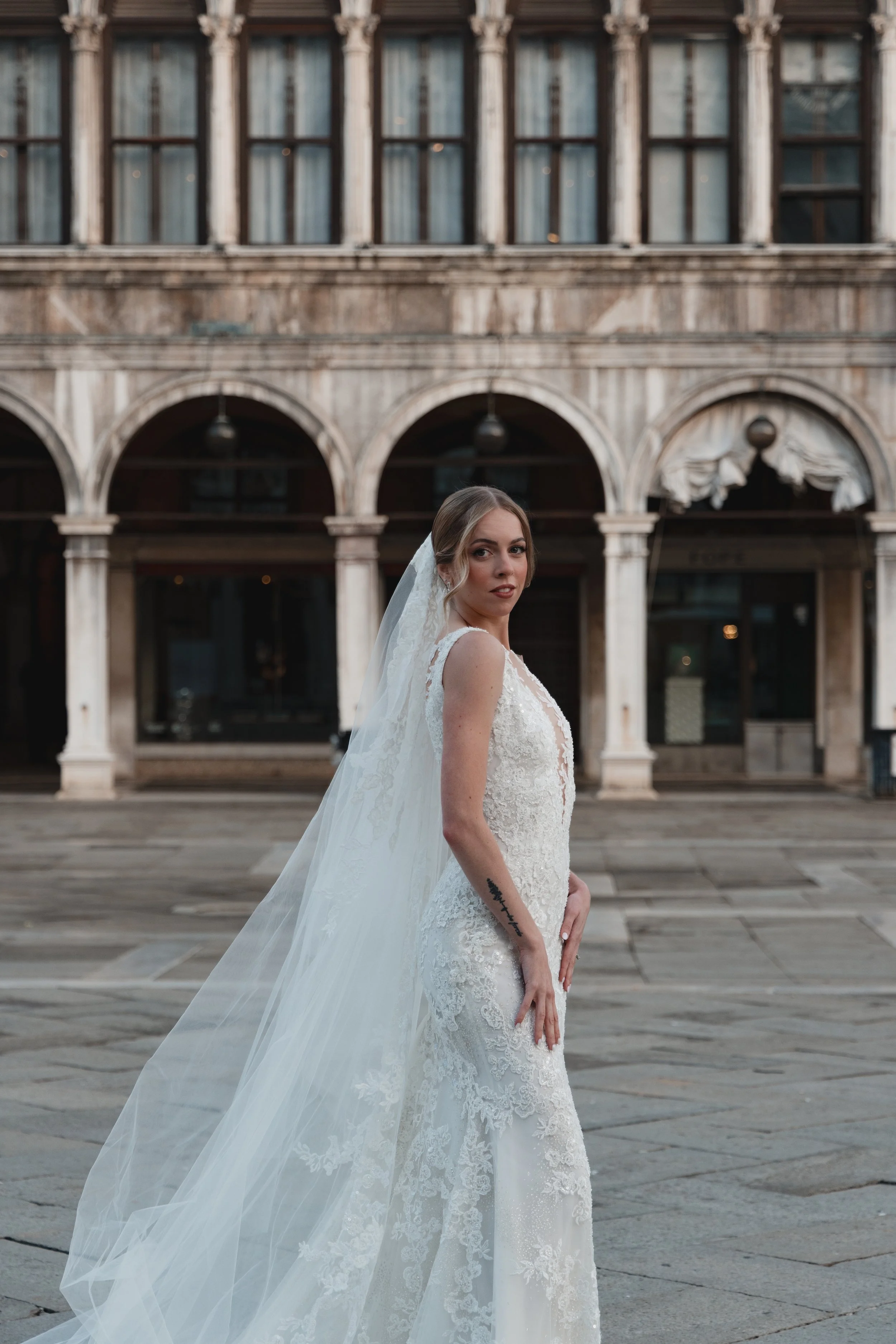 A bride in a white lace wedding gown with a veil stands in front of an old stone building with arches and tall windows.
