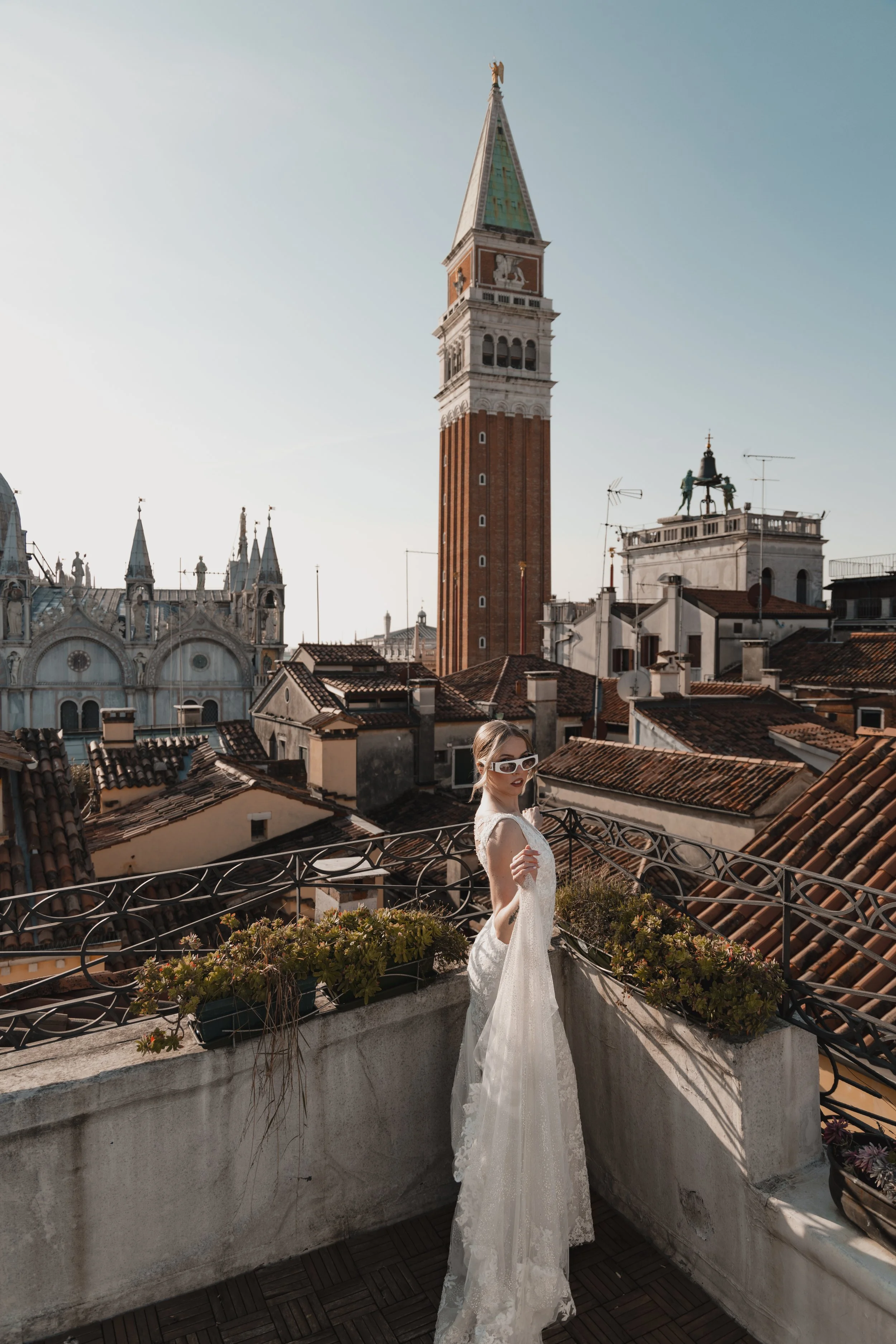 Woman in a white wedding dress and sunglasses standing on a rooftop balcony with historic European buildings and the clock tower of St. Mark's Campanile in Venice in the background.