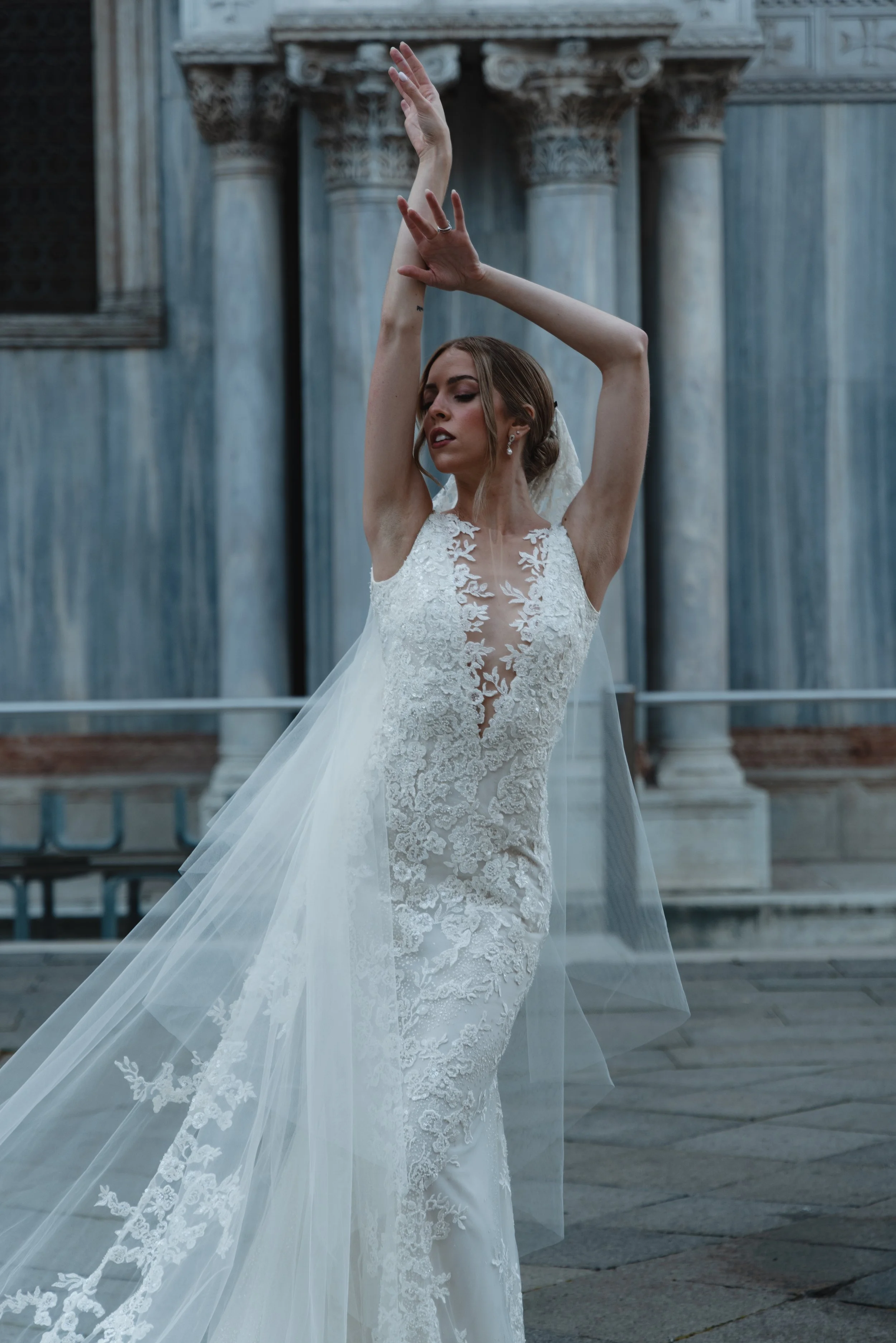 A woman in a white wedding dress with lace details standing in front of a blue marble building with classical columns and raised hands in a graceful pose.