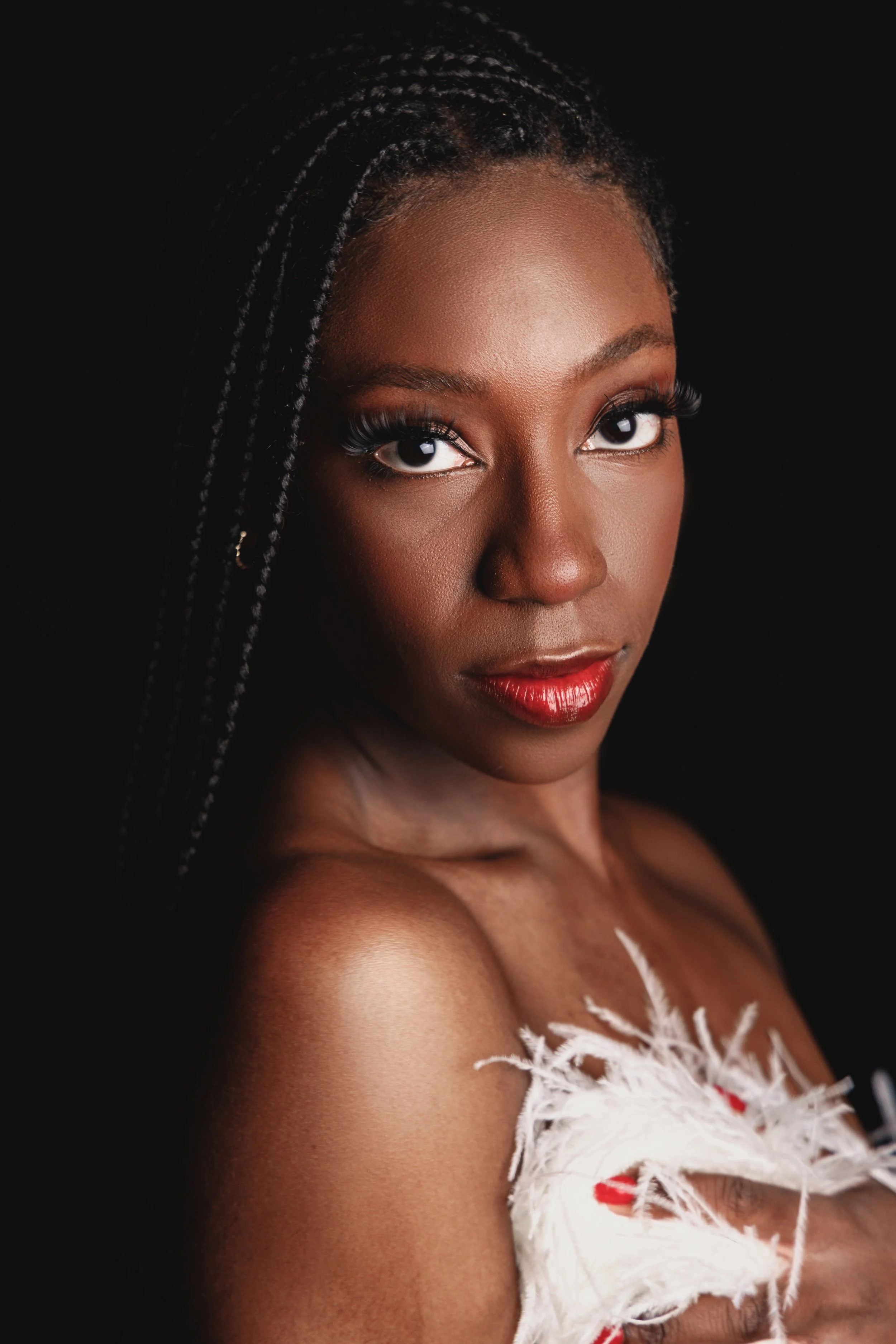 Close-up portrait of a woman with dark skin, styled with long braided hair, makeup including bright red lipstick, and dressed in a white feathered garment against a black background.
