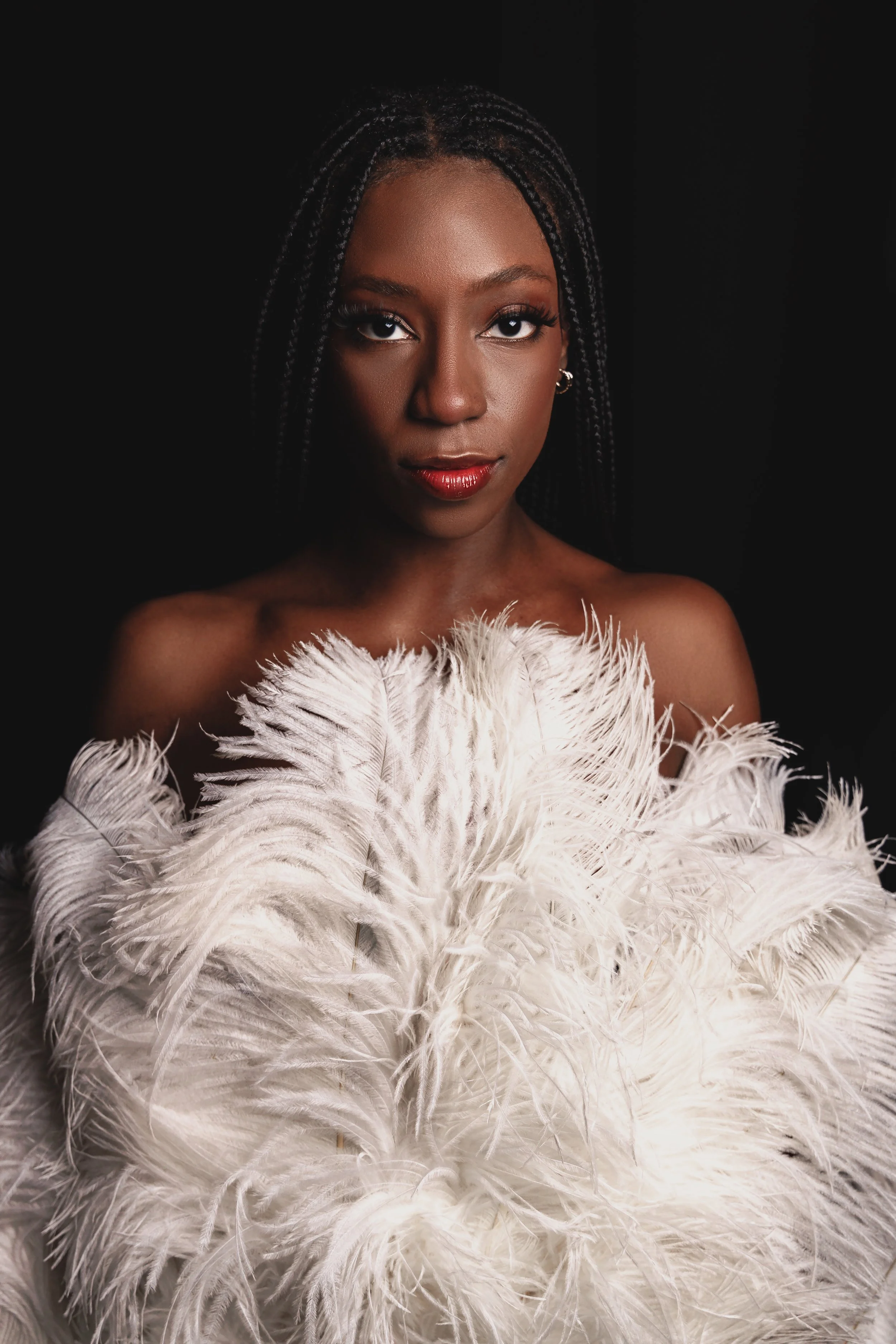 A woman with dark skin tone and braided hair stands against a black background. She wears a white feathered garment or accessory around her shoulders.