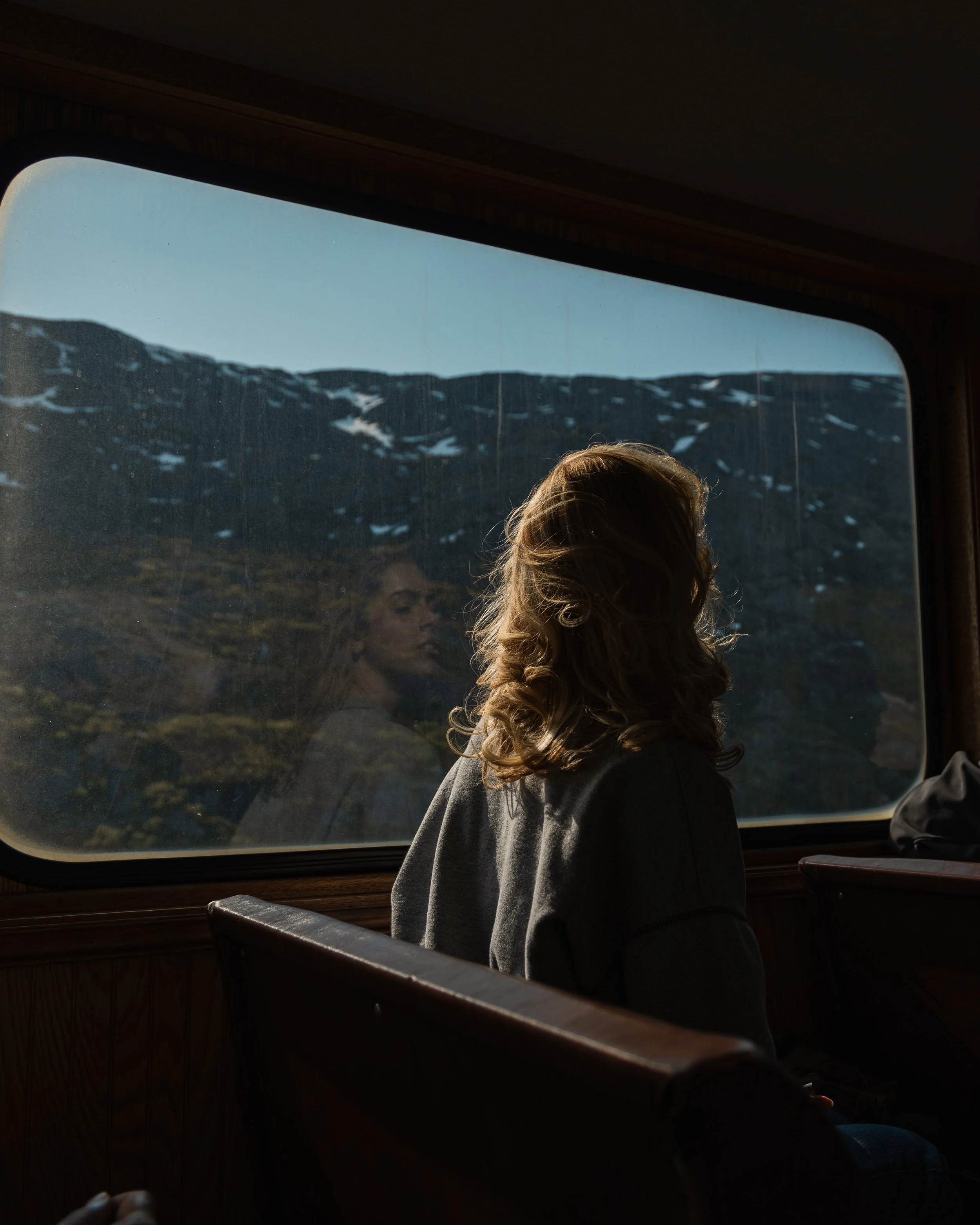 Inside a moving train, showing a woman with curly blond hair looking out the window at snow-covered mountains and a clear sky.