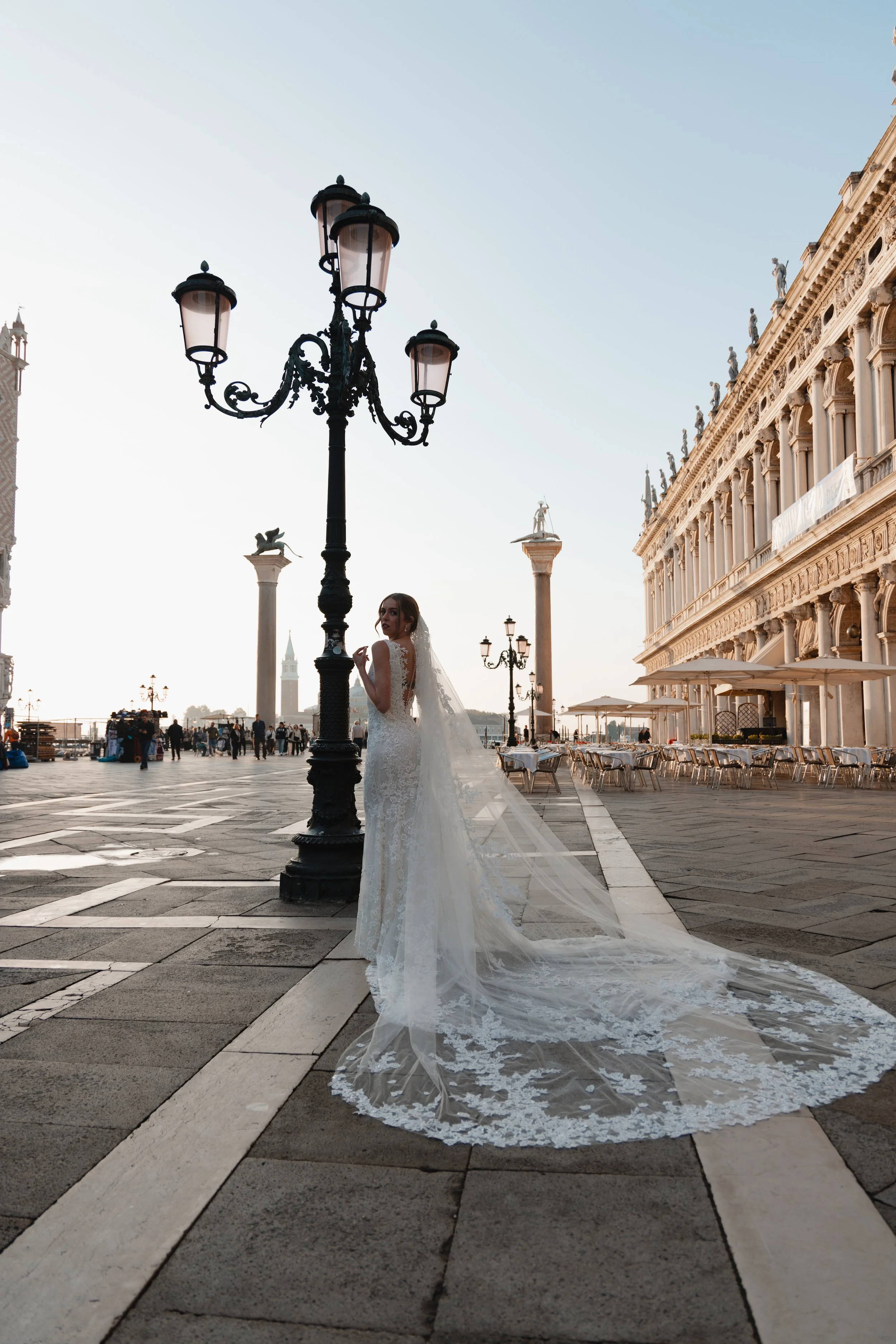 A bride in a white wedding gown with long train standing on a city square at sunset, next to a vintage black lamppost and historic buildings with statues on the roof. Outdoor seating and people are in the background.