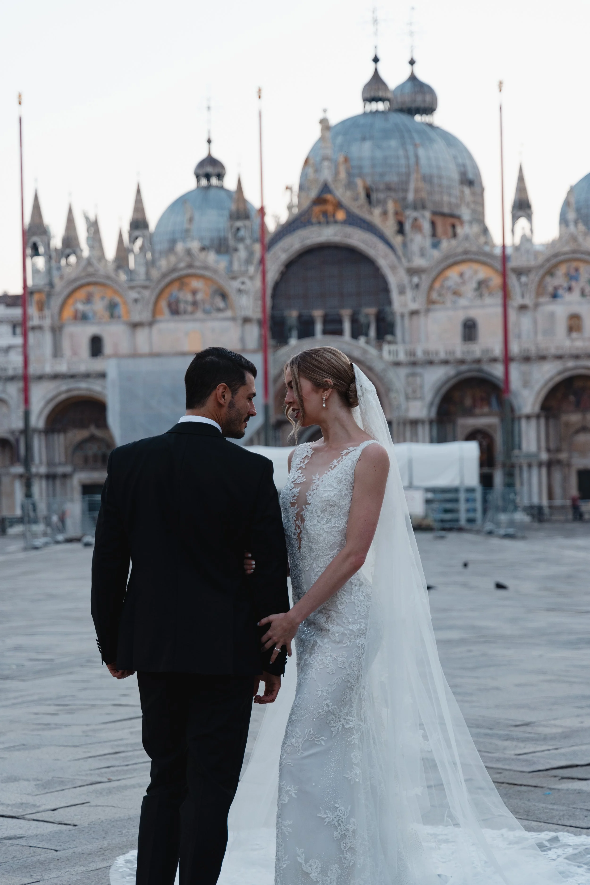 A bride and groom holding hands, facing each other, in front of St. Mark's Basilica in Venice, Italy, during their wedding.
