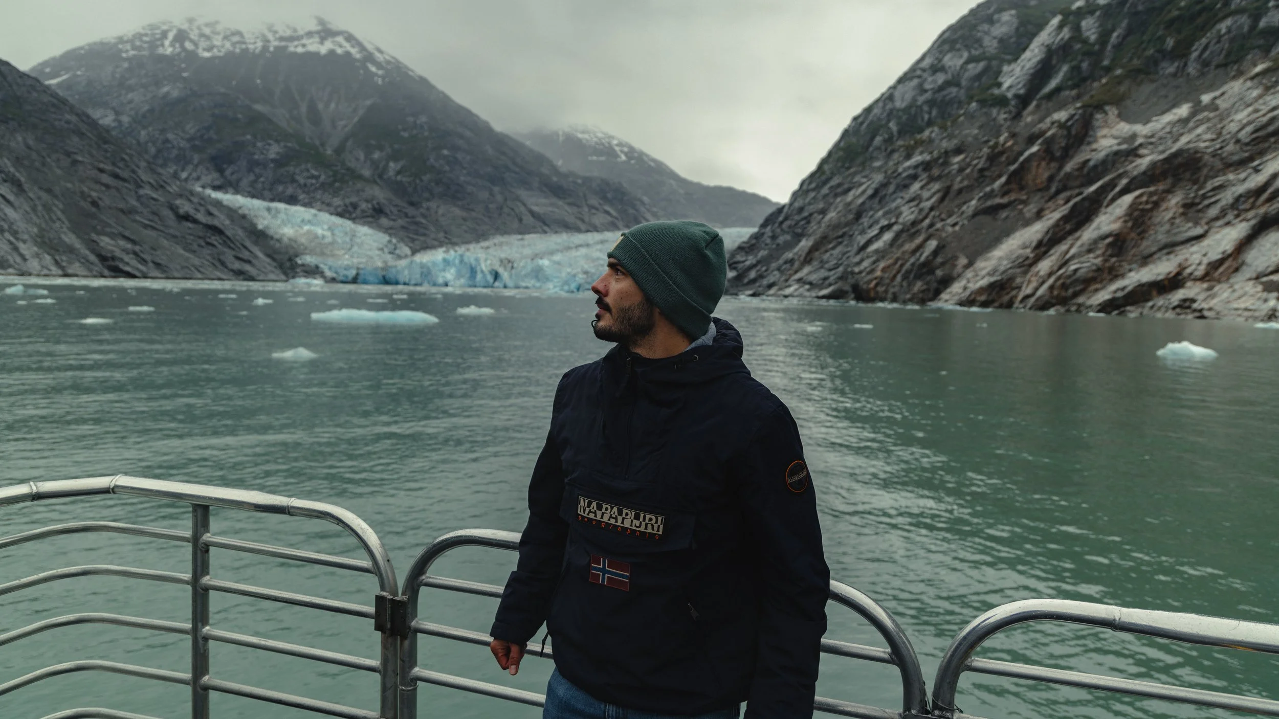 A man wearing a green beanie and black jacket standing on a boat in a fjord with mountains and a glacier in the background.