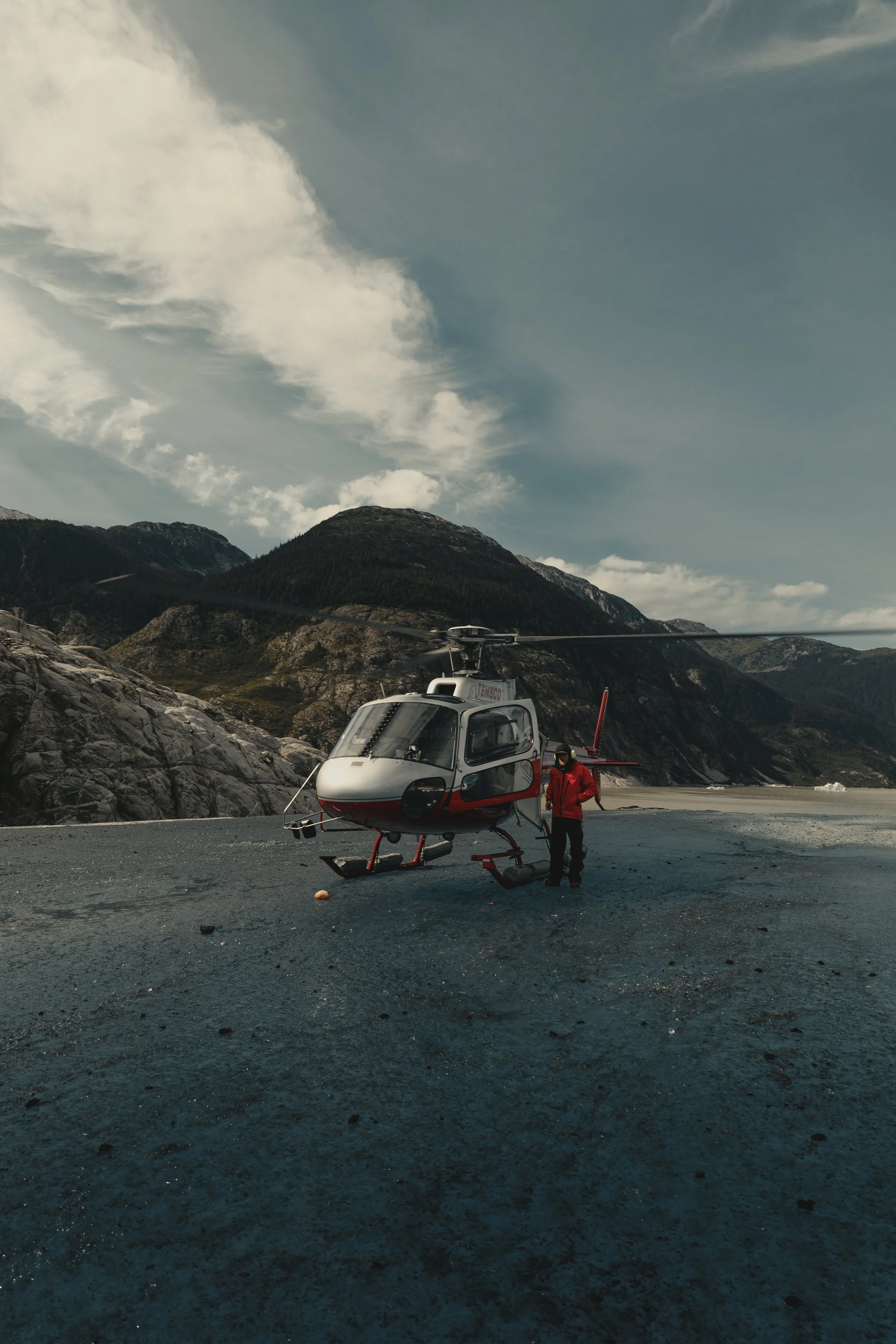 A helicopter on a rocky terrain near a mountainous landscape under a partly cloudy sky, with a person in a red jacket standing beside it.