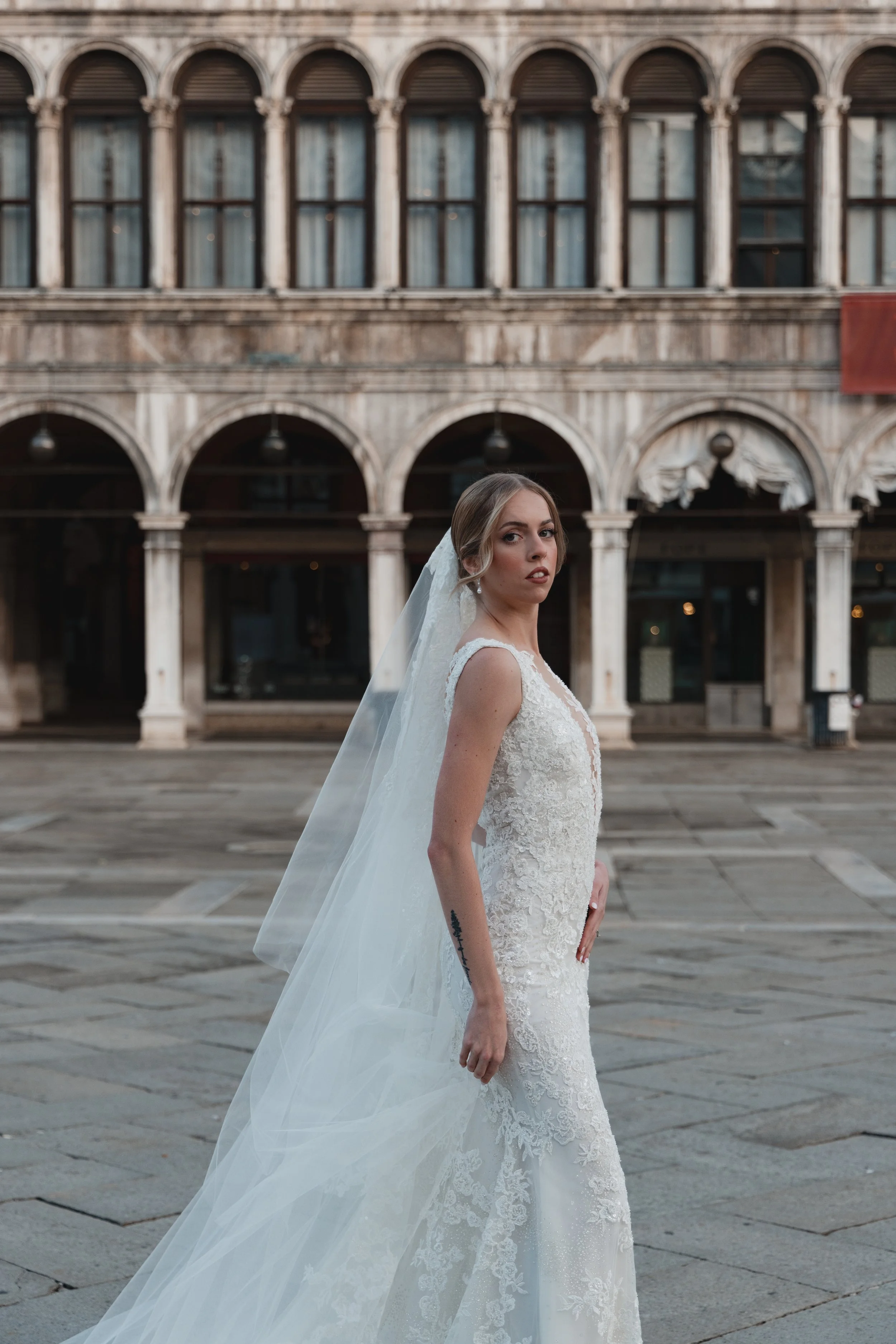 A woman in a white wedding dress and veil standing outside in front of a historic building with arches and columns.