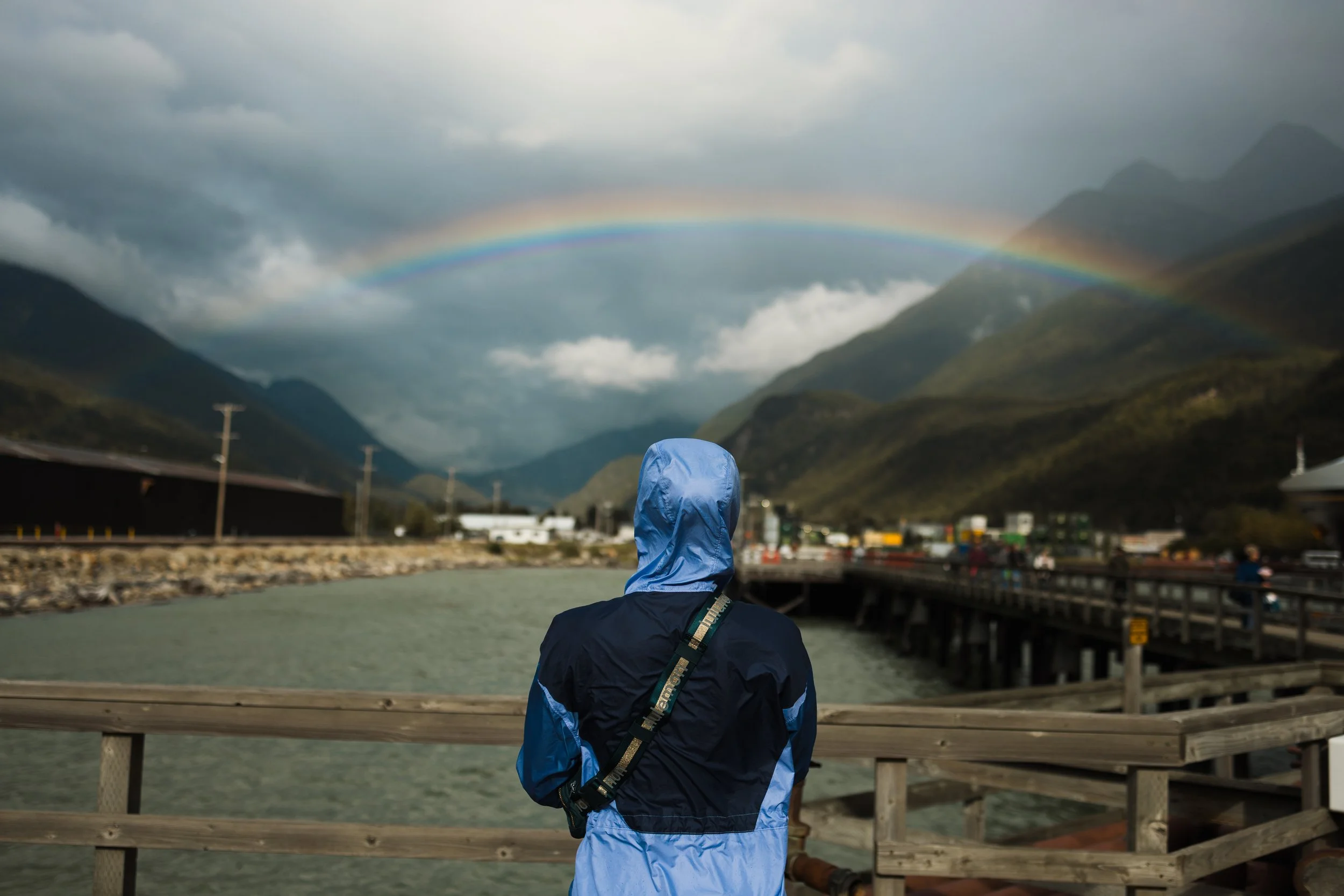 A person wearing a blue raincoat standing on a wooden bridge, looking at a double rainbow in the cloudy sky above mountains and buildings.