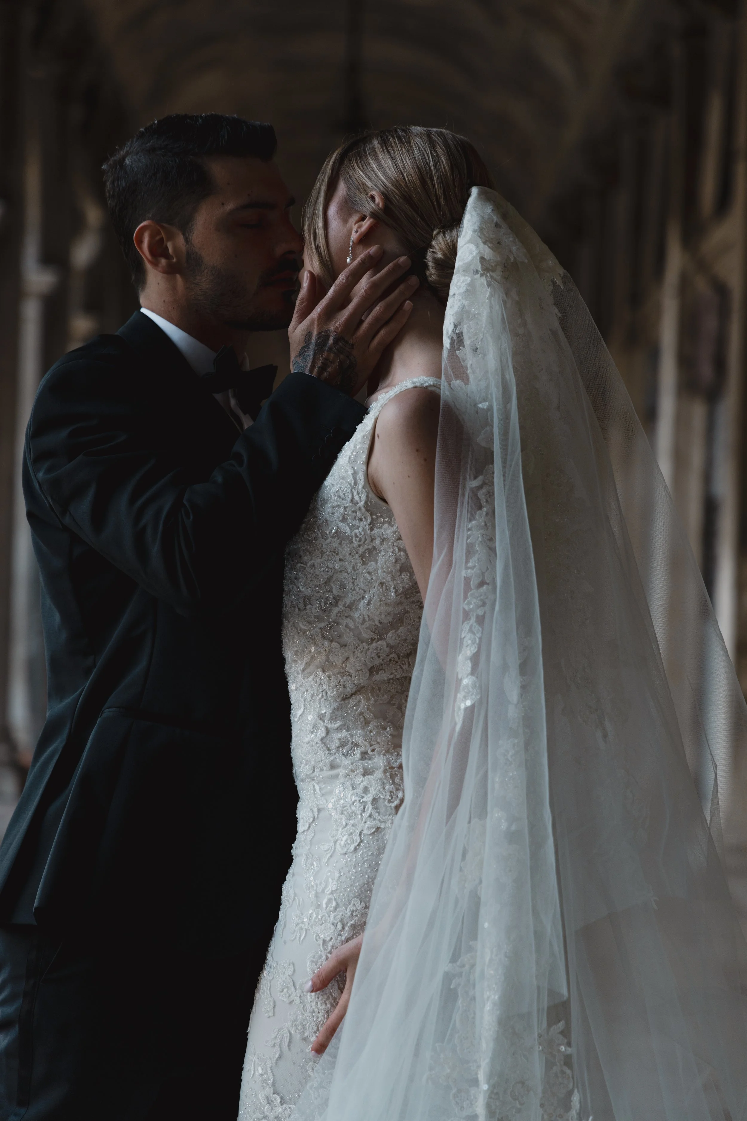 A groom in a black tuxedo and a bride in a lace wedding dress and veil sharing an intimate moment with closed eyes and touching faces inside a grand stone building.