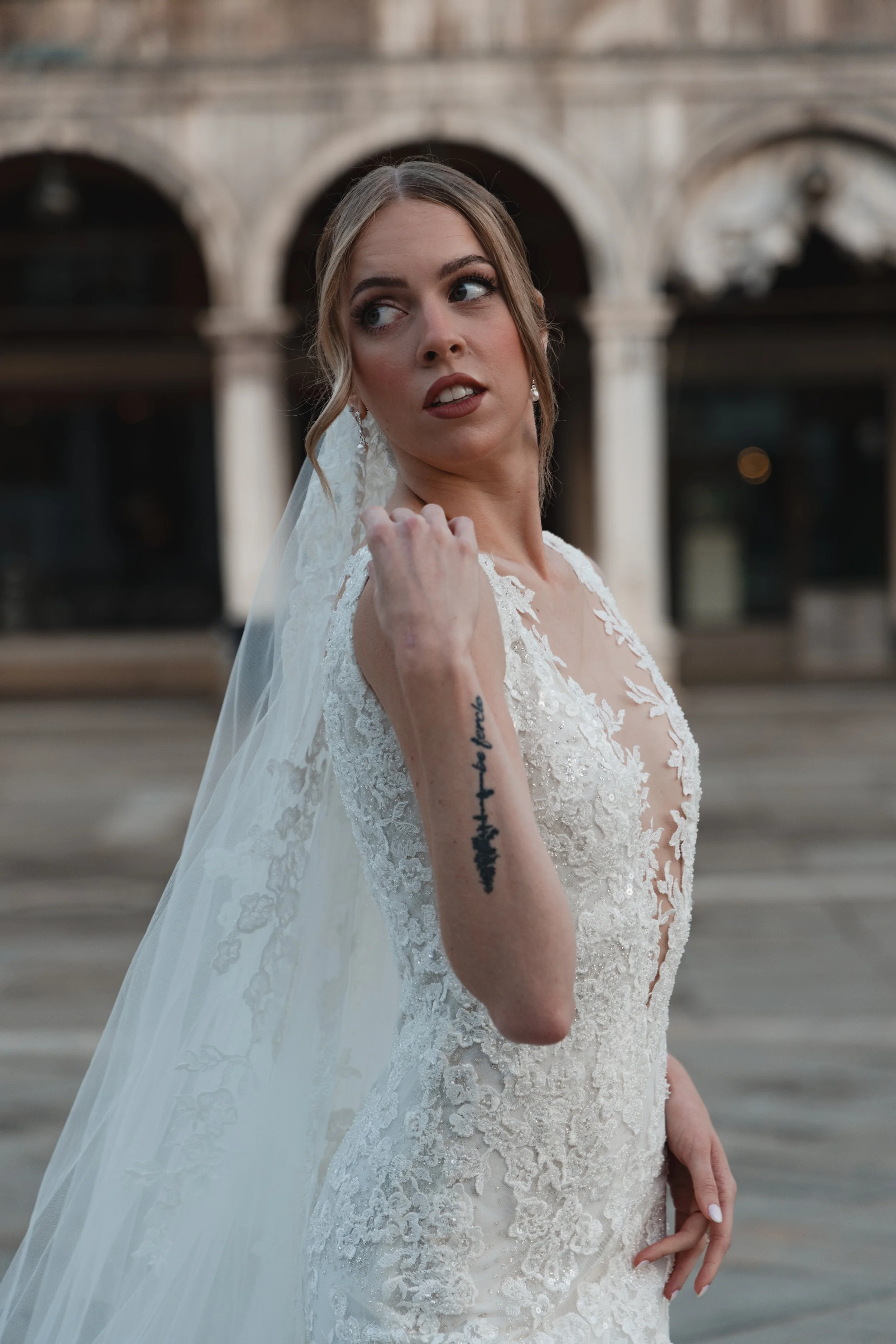 A woman in a white lace wedding dress with a veil, looking over her shoulder, standing outdoors near an arched stone background.