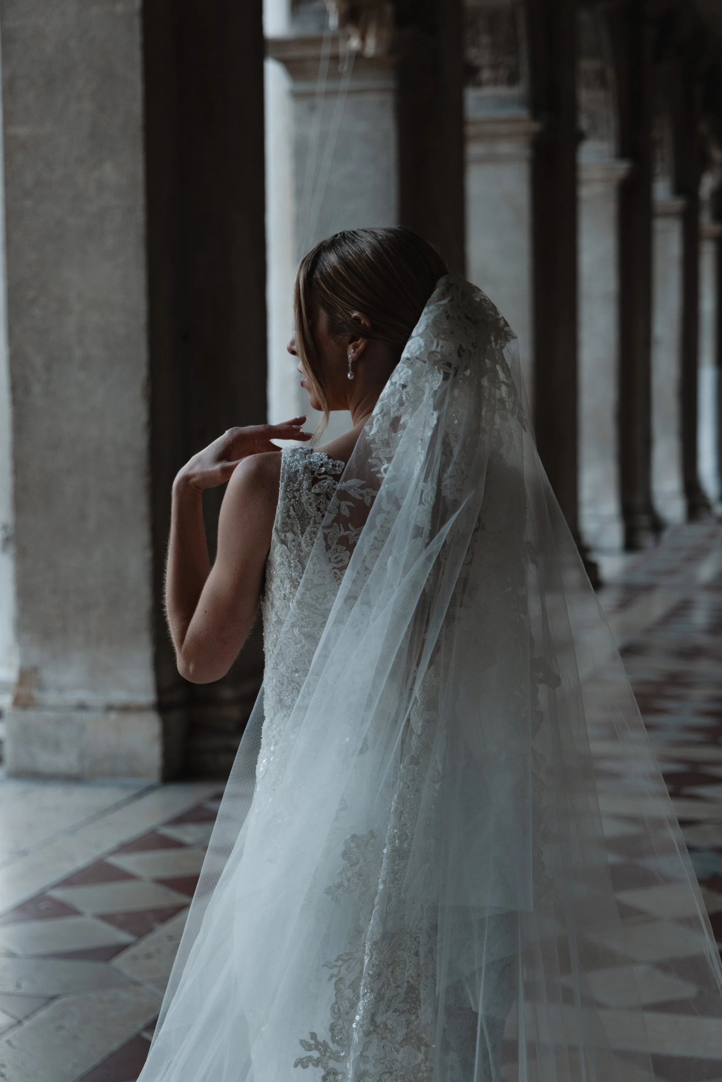 A bride in a wedding dress with lace details and a veil, standing in a historic building with stone columns and checkered floor tiles.