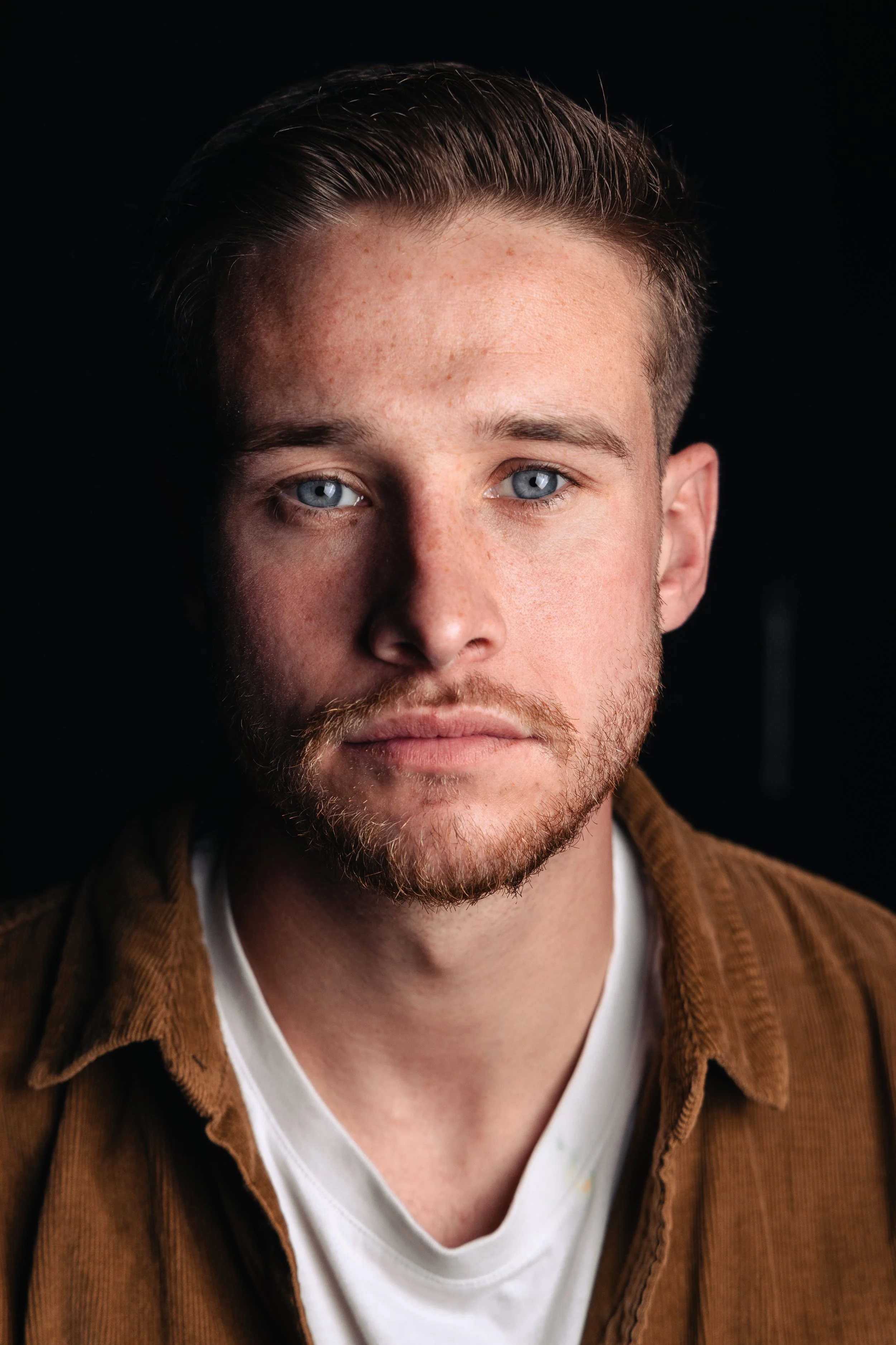Close-up of a young man with blue eyes, light brown hair, and a beard, wearing a white shirt and a brown jacket, against a black background.