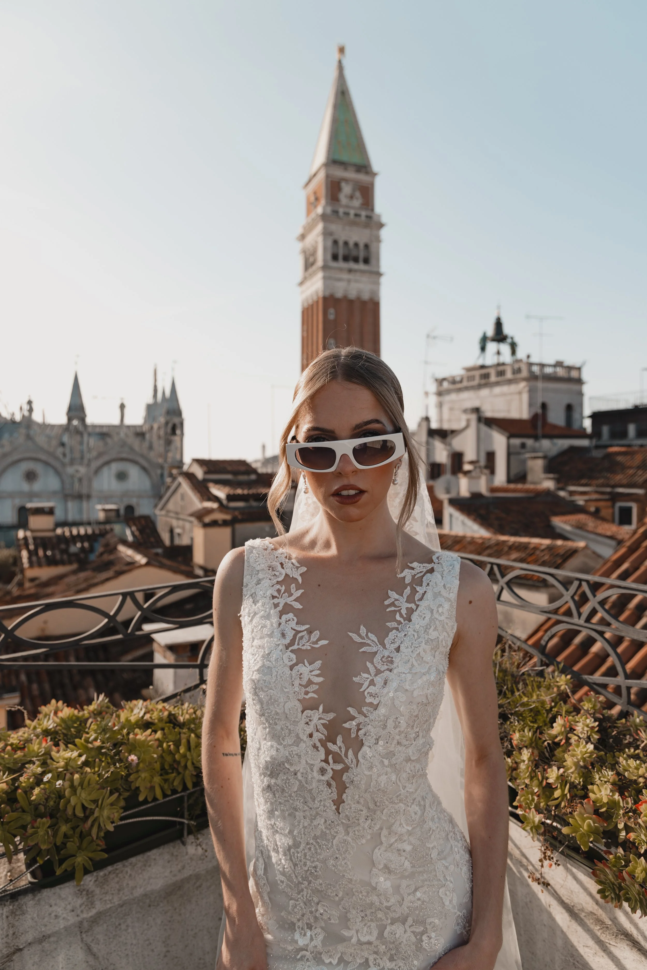 A bride in a white lace wedding dress and large white sunglasses stands on a rooftop with the Venice cityscape in the background, including the famous bell tower and rooftops of Venice, Italy.