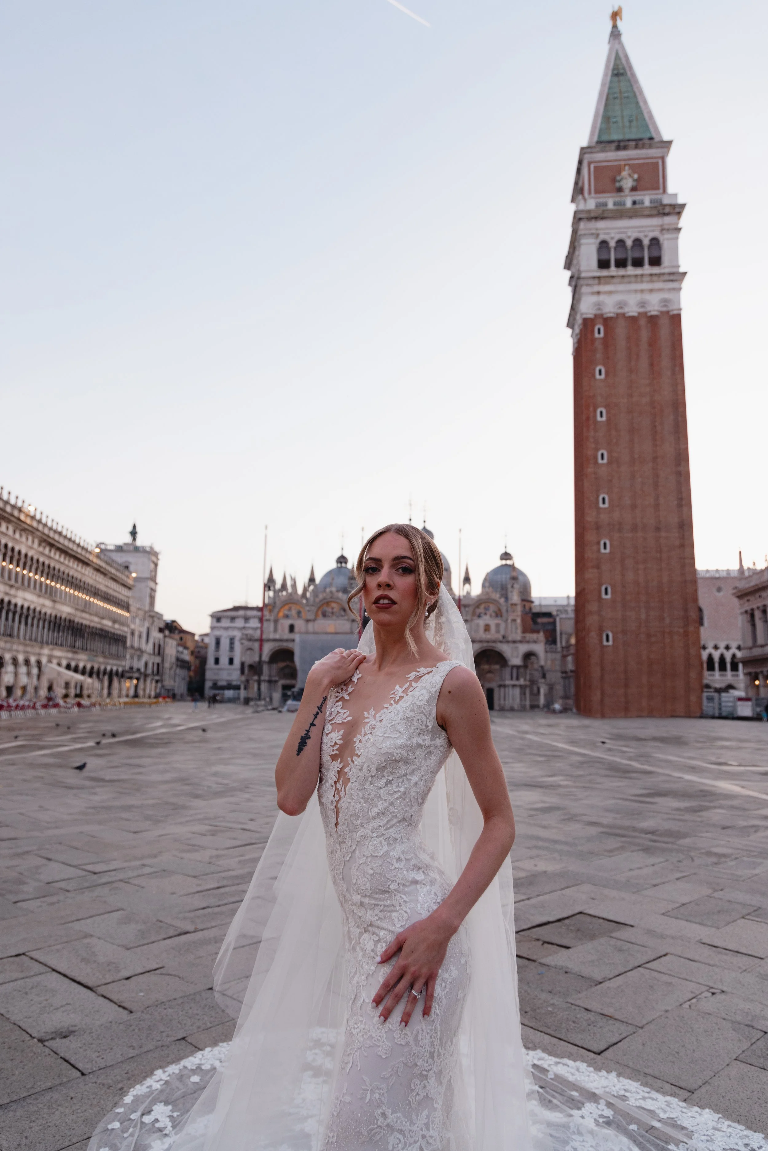 A woman in a wedding dress standing in Piazza San Marco, Venice, with the Campanile di San Marco in the background during dusk.