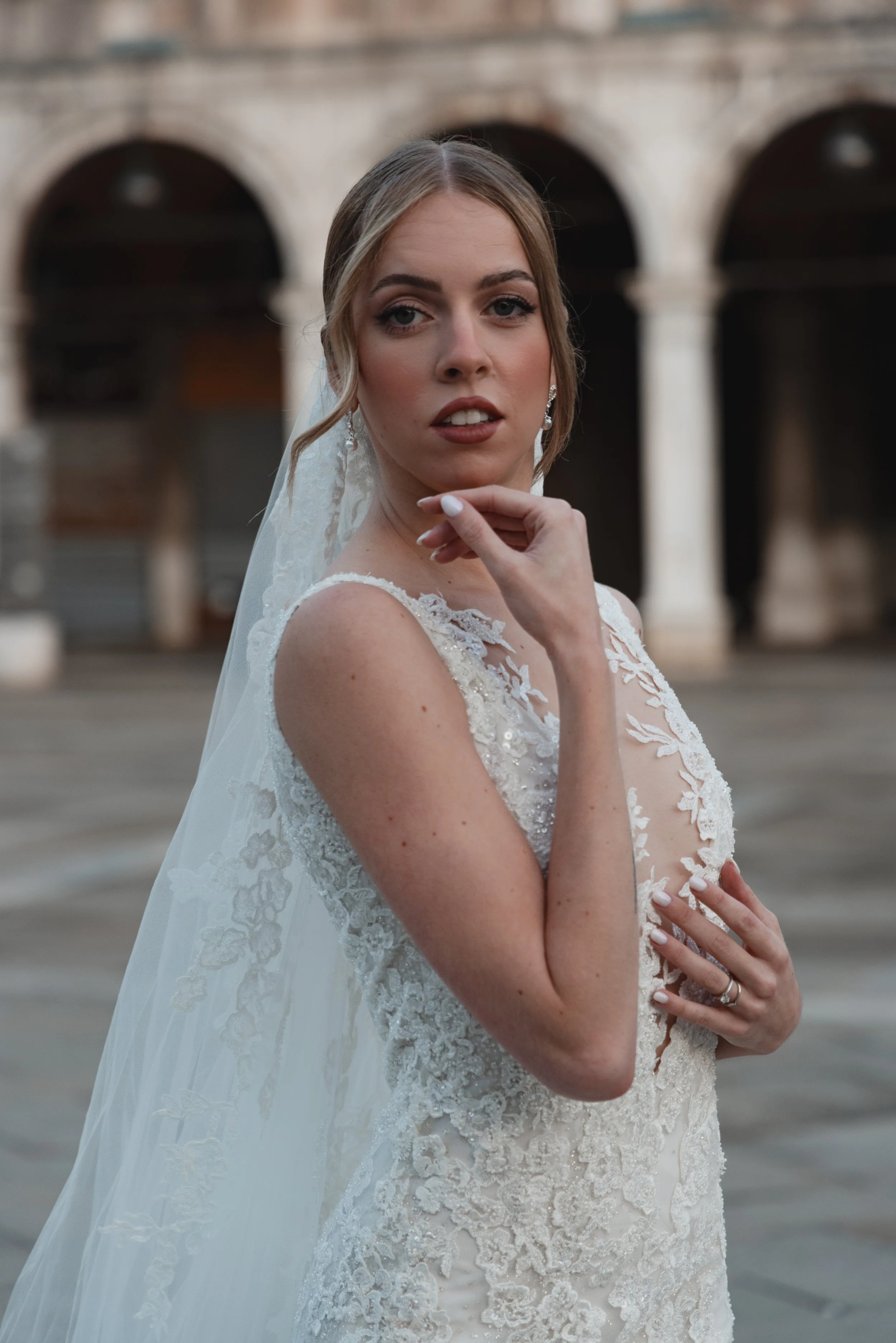 Close-up of a bride in a lace wedding dress with a veil, touching her chin with her right hand, in front of an arched brick structure.
