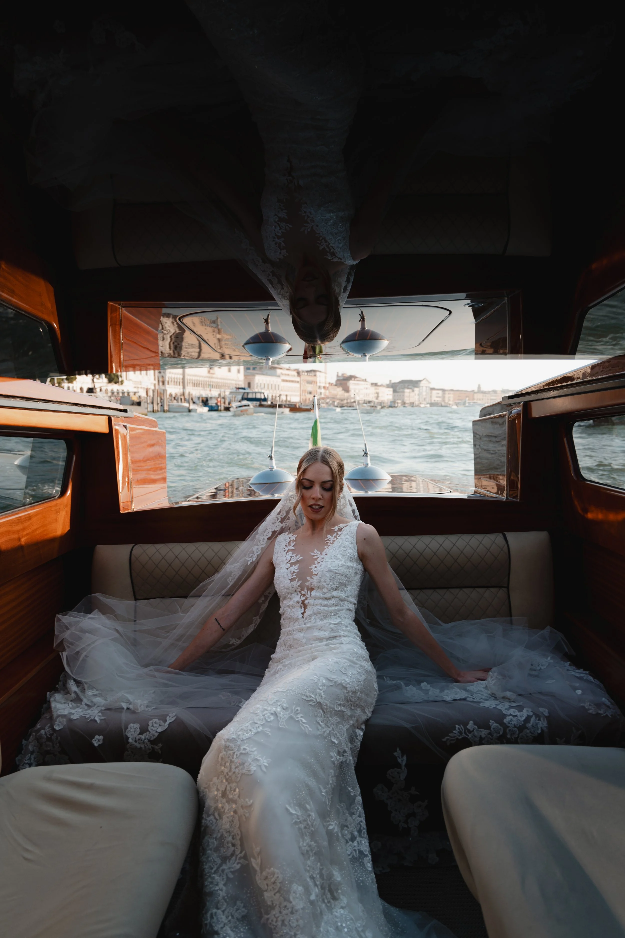 A bride dressed in a white lace wedding gown sitting inside a boat with a city and waterway visible in the background.