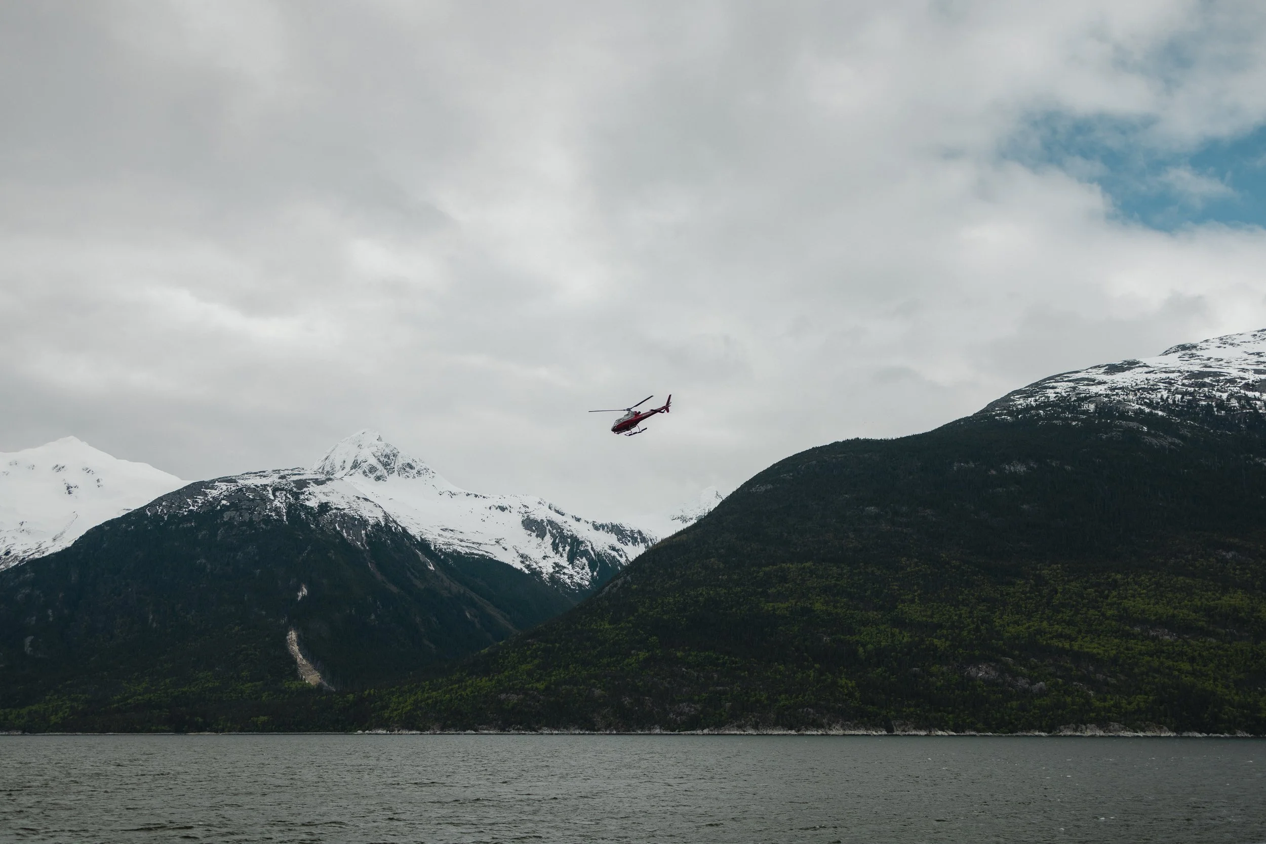 A helicopter flying over a mountainous landscape with snow-capped peaks, green forests, and a body of water in the foreground under cloudy skies.