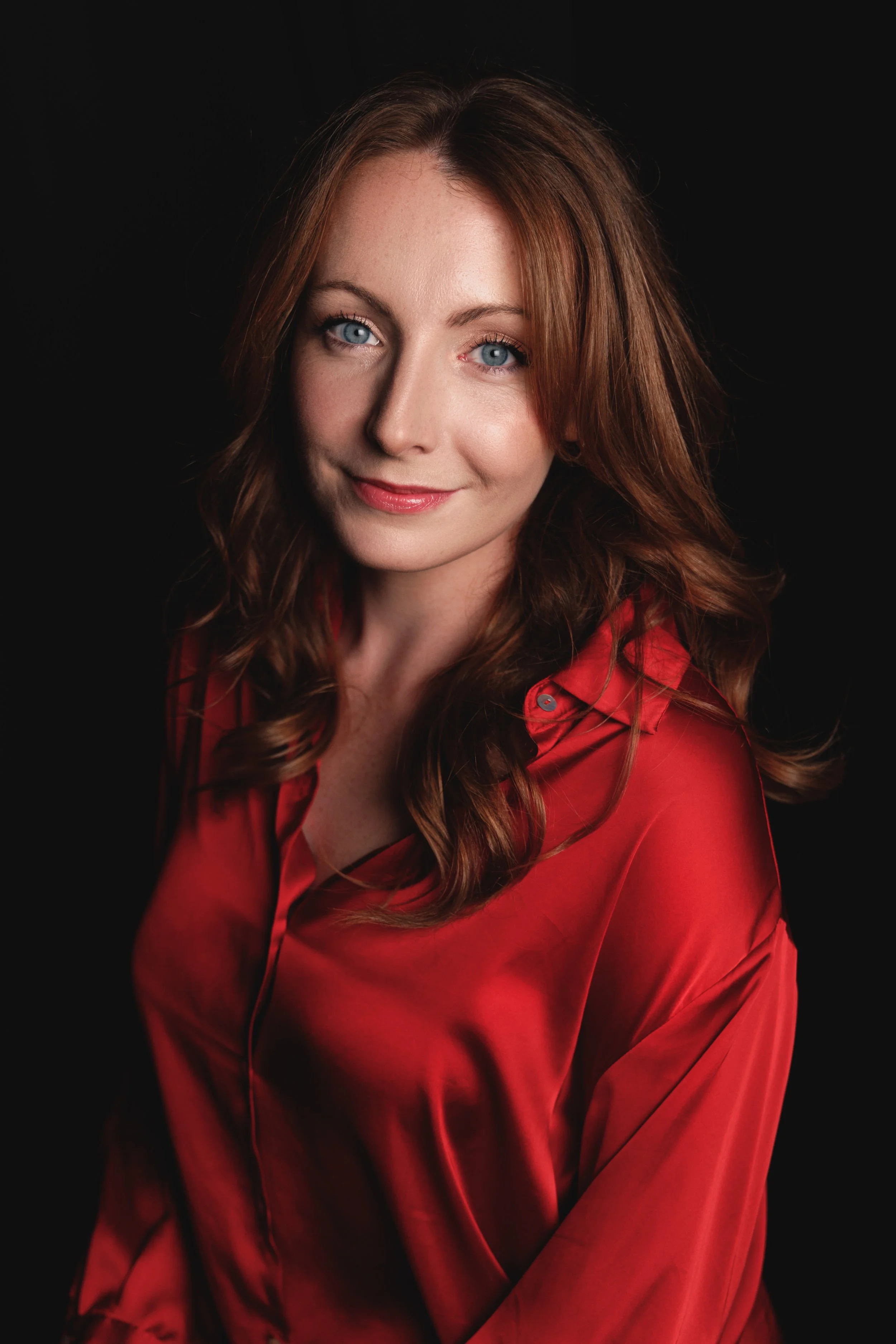 A woman with red hair and blue eyes wearing a red satin shirt against a black background.