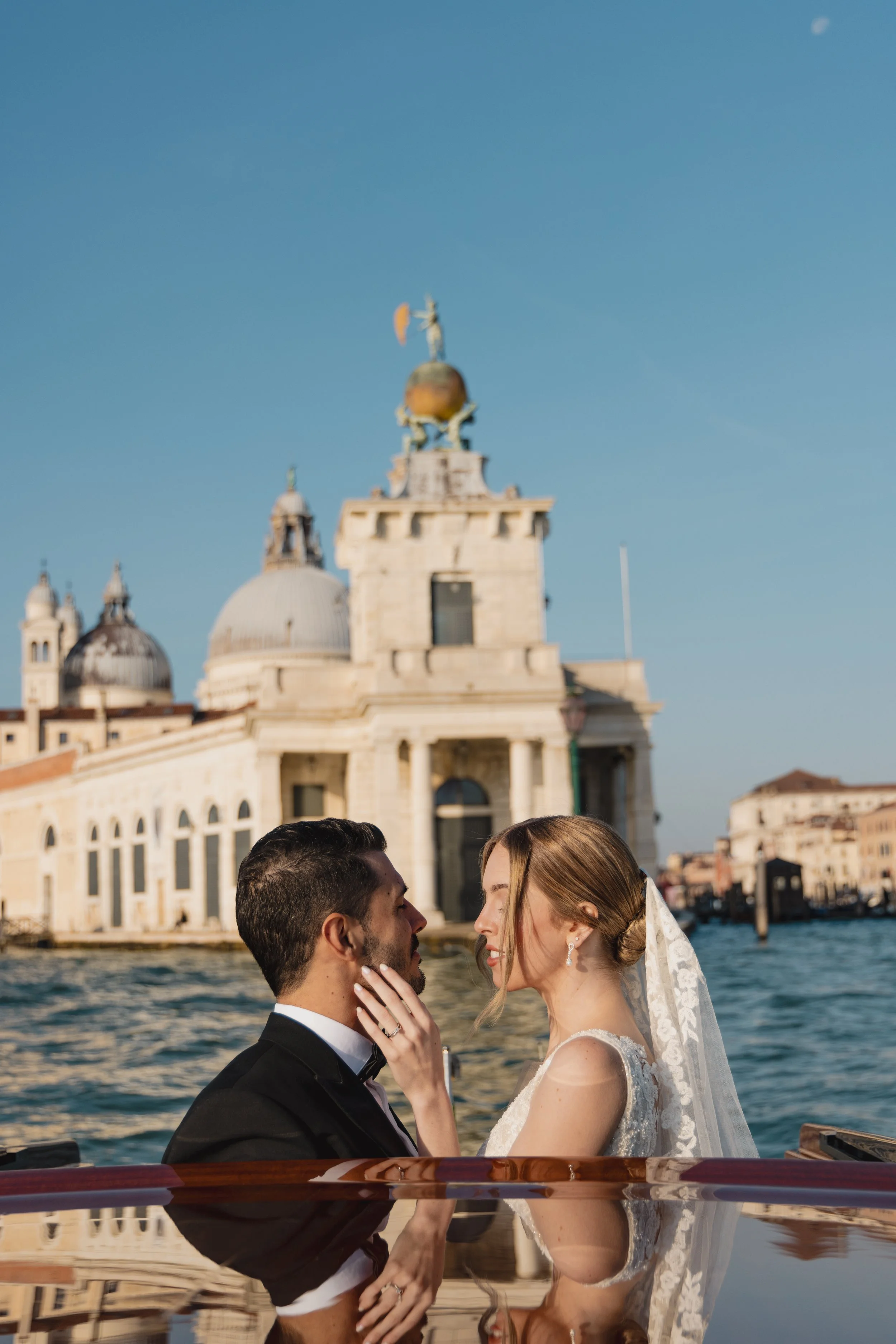 A couple dressed in wedding attire, close together on a boat in Venice, Italy, with historic buildings and water in the background.