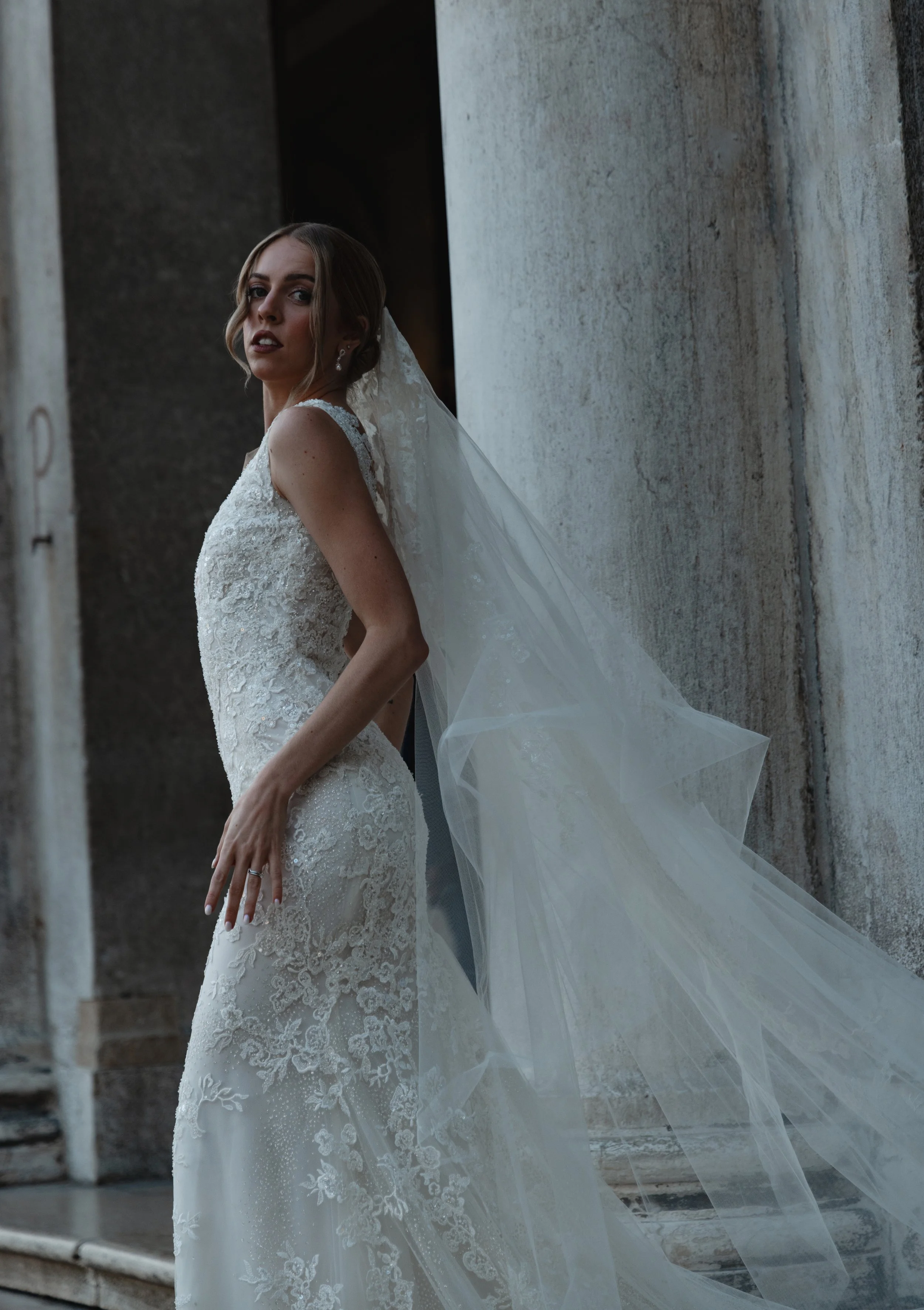 A bride in a lace wedding dress with a long veil poses against a concrete wall.
