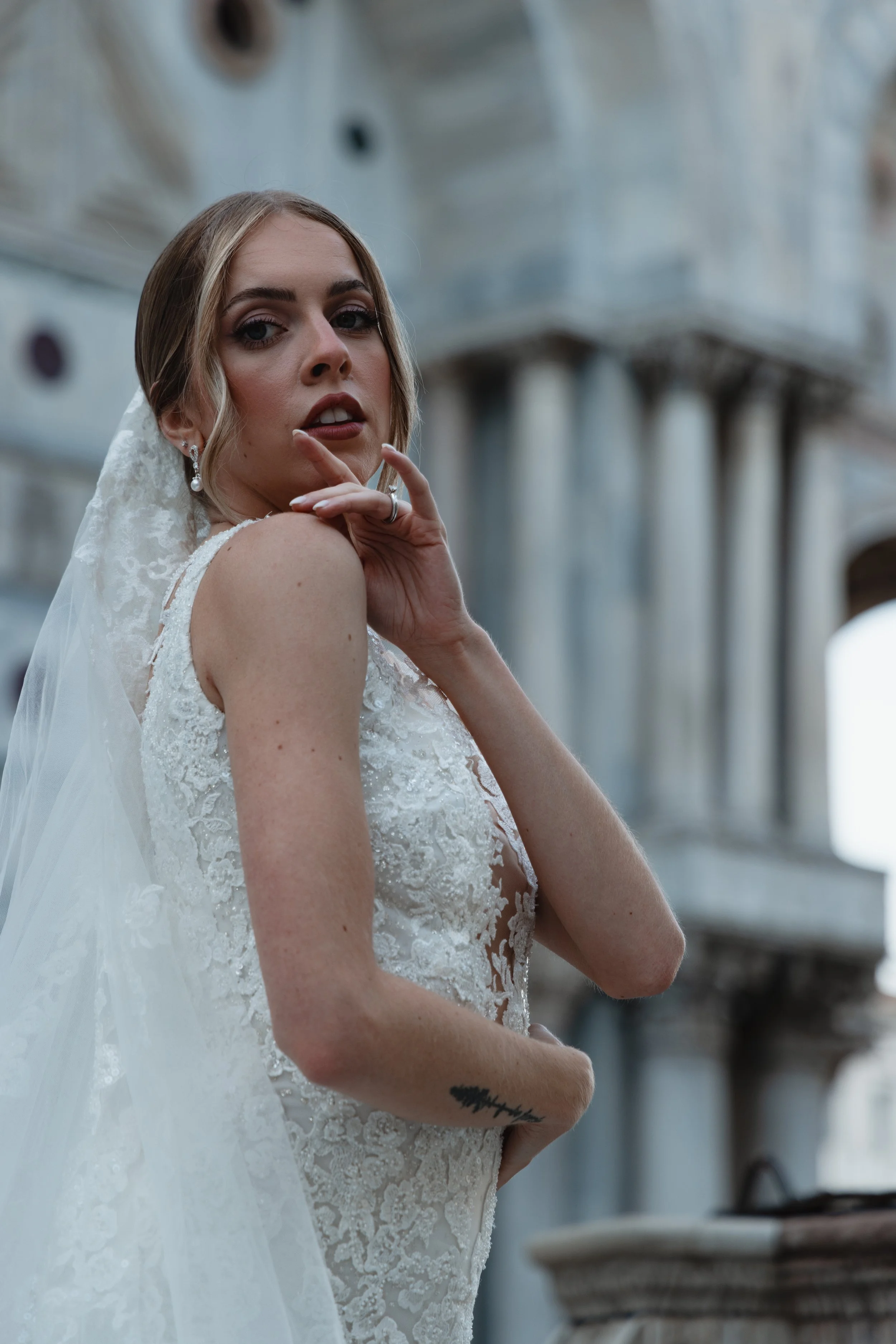 A bride with light brown hair in soft curls, wearing a white lace wedding dress and pearl earrings, posing in front of a light-colored building with architectural details.