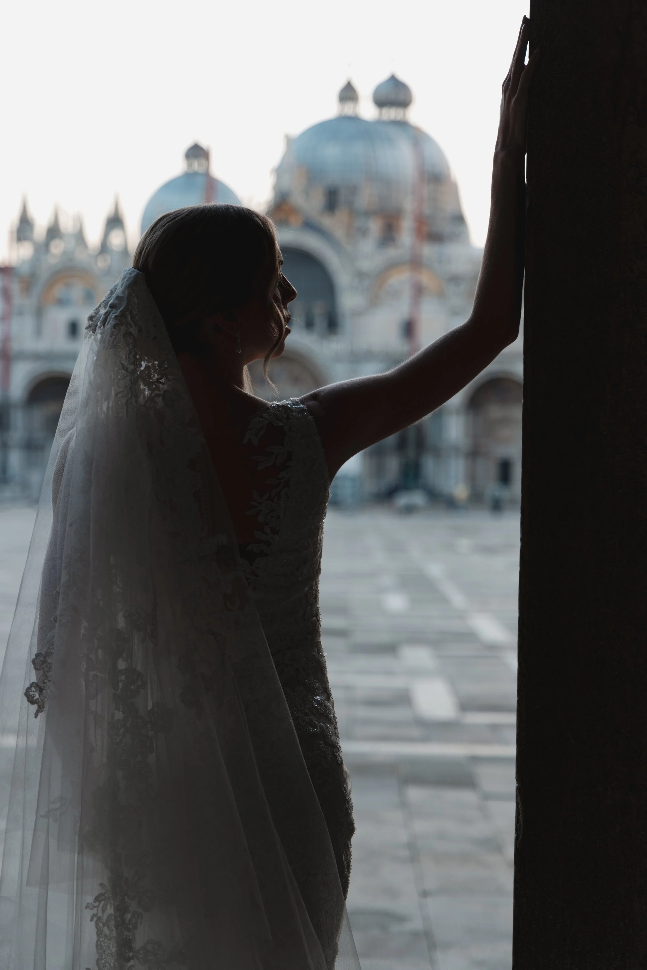 A silhouette of a woman in a wedding dress with a veil, touching a dark doorway or wall, with a blurred historic European-style building with domes in the background.