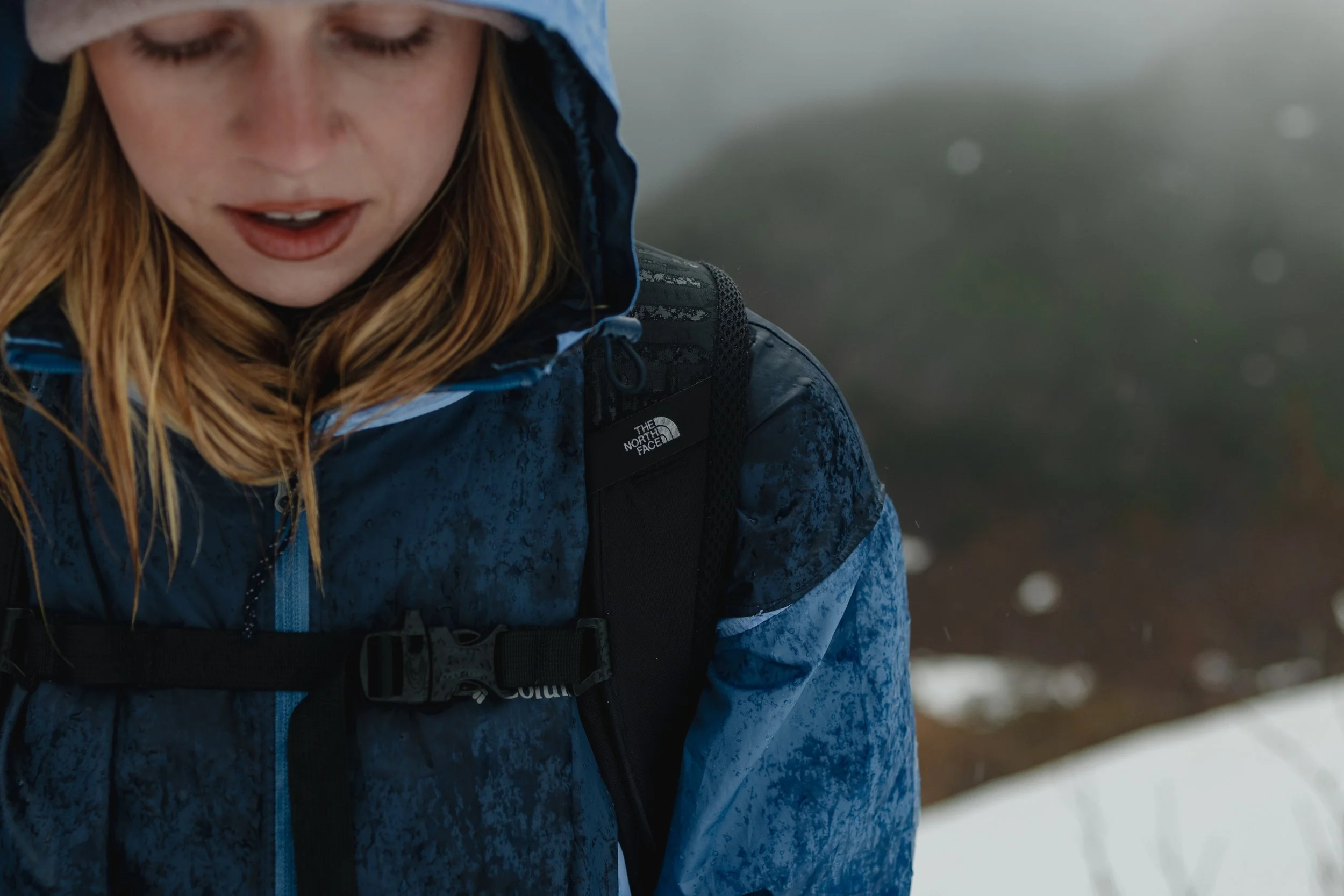 Close-up of a woman with wet red hair wearing a blue waterproof jacket and a black backpack outdoors in snowy weather.