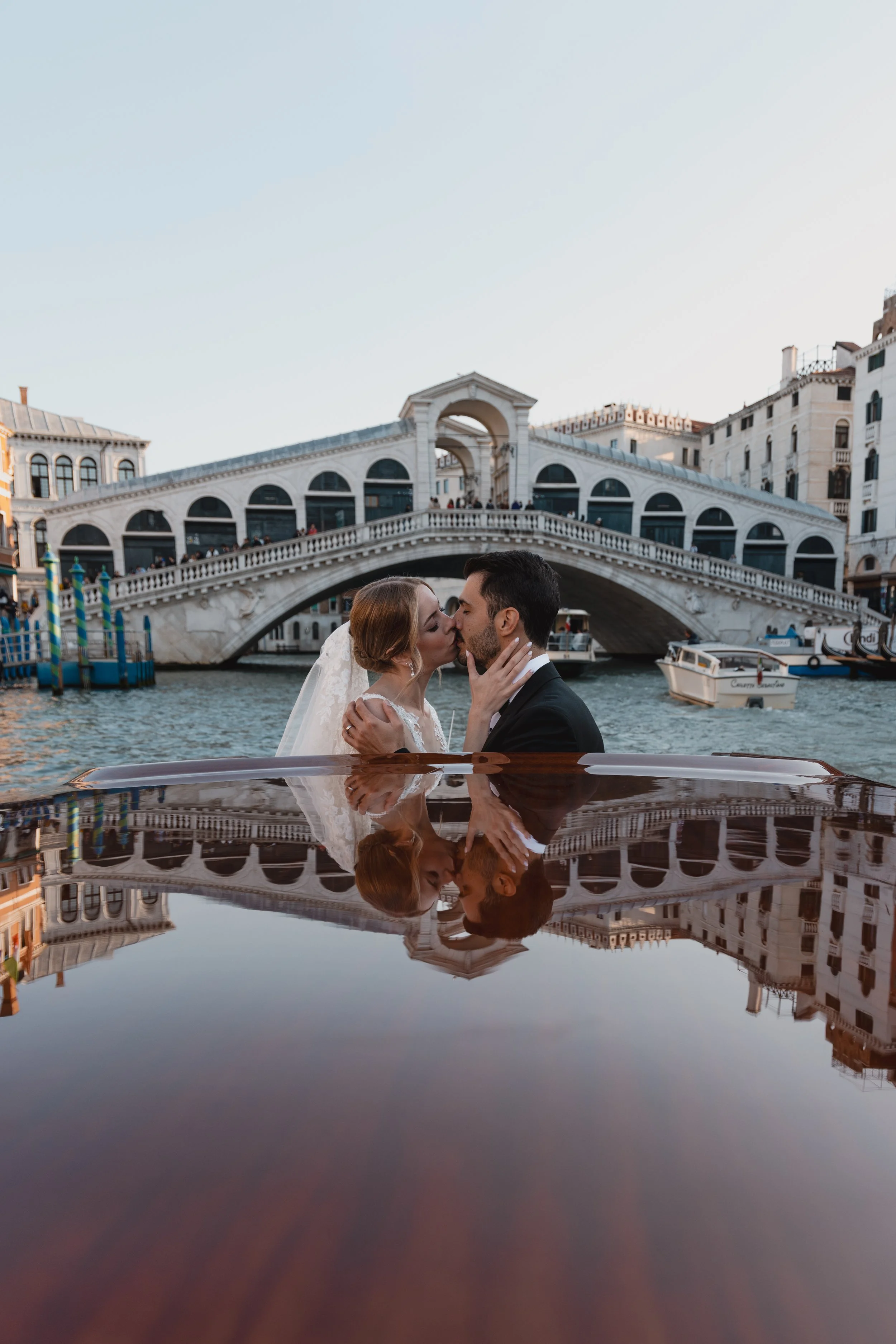 A bride and groom kissing on a boat in front of a historic bridge with buildings in Venice, Italy, during sunset.
