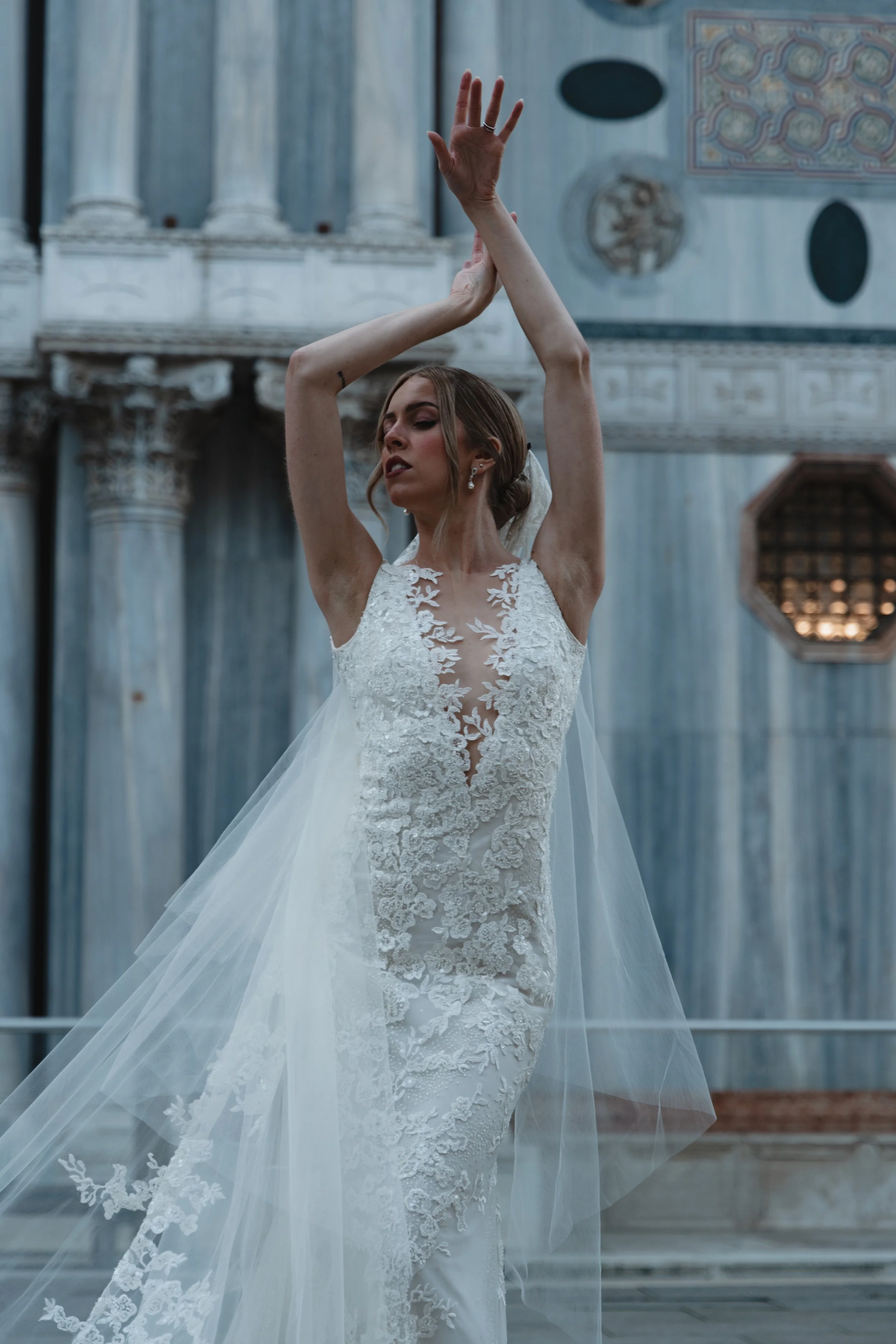 A woman in a wedding dress standing outdoors, with her eyes closed and arms raised above her head in a dance pose. The background features classical architectural elements and artwork on a building.