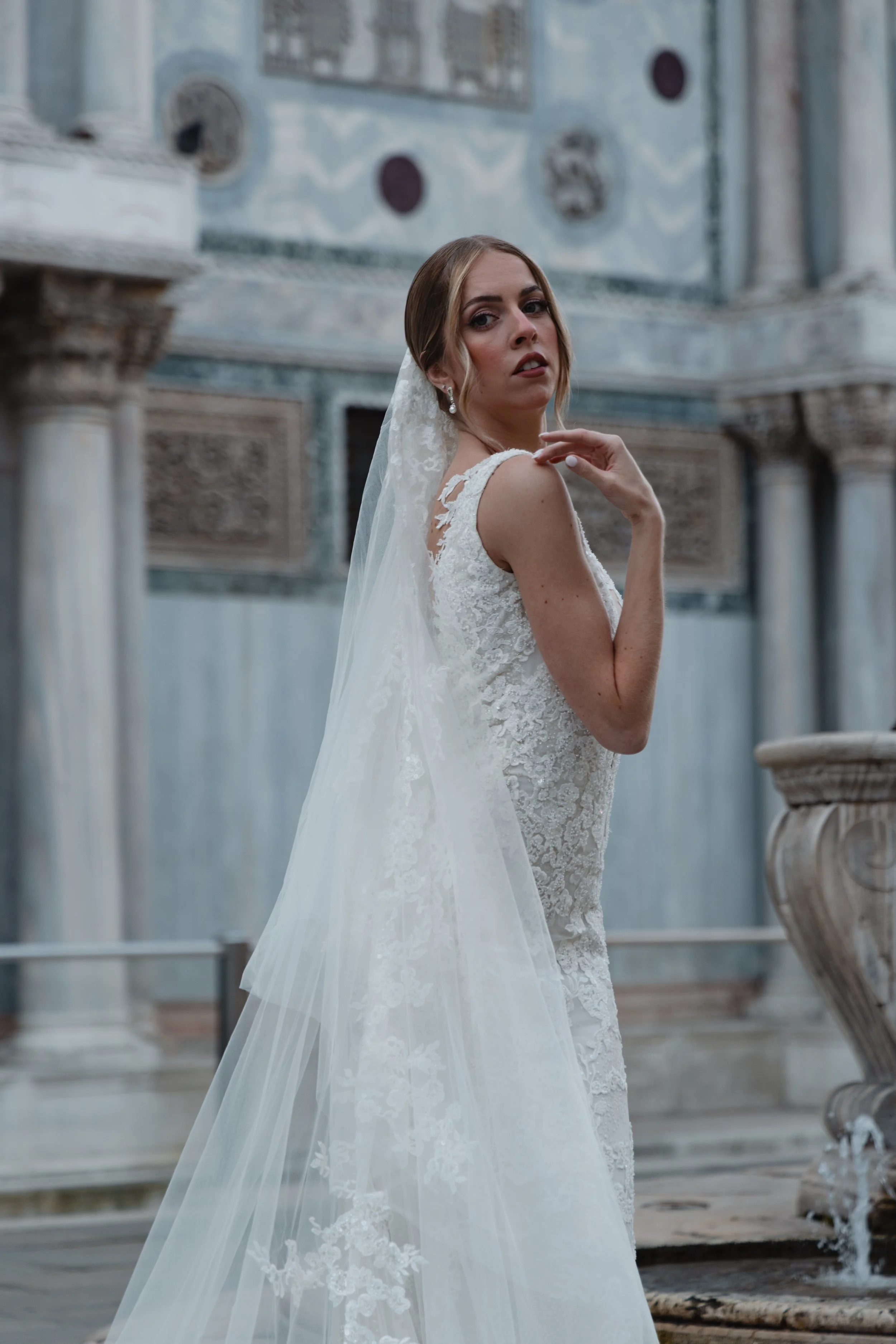 A bride in a white lace wedding gown with a sheer train, wearing pearl earrings, standing outdoors near a marble fountain.