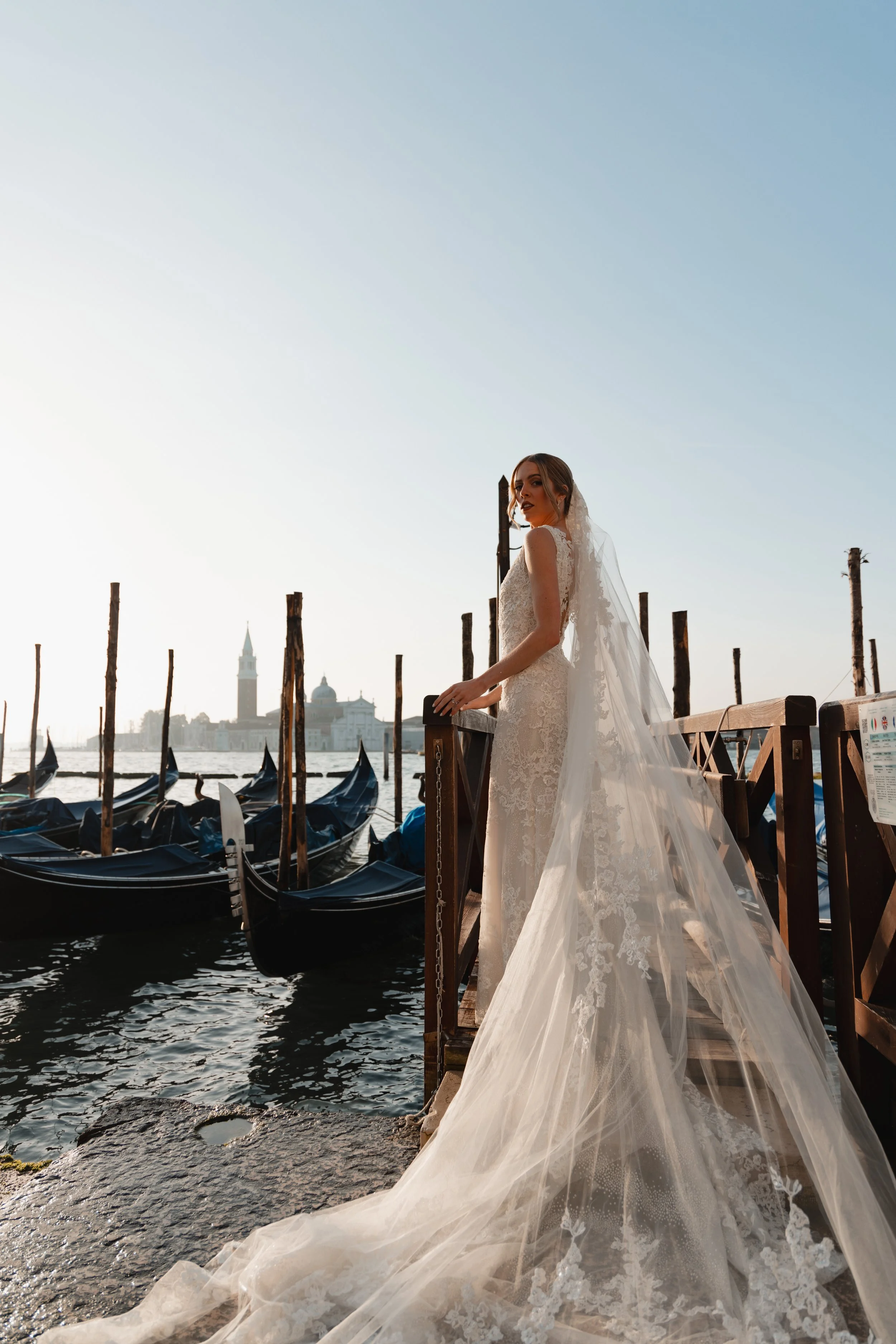 A bride in a white wedding dress with lace details and a long train stands on a dock by the water, with gondolas docked nearby and the city of Venice, Italy, in the background during daylight.