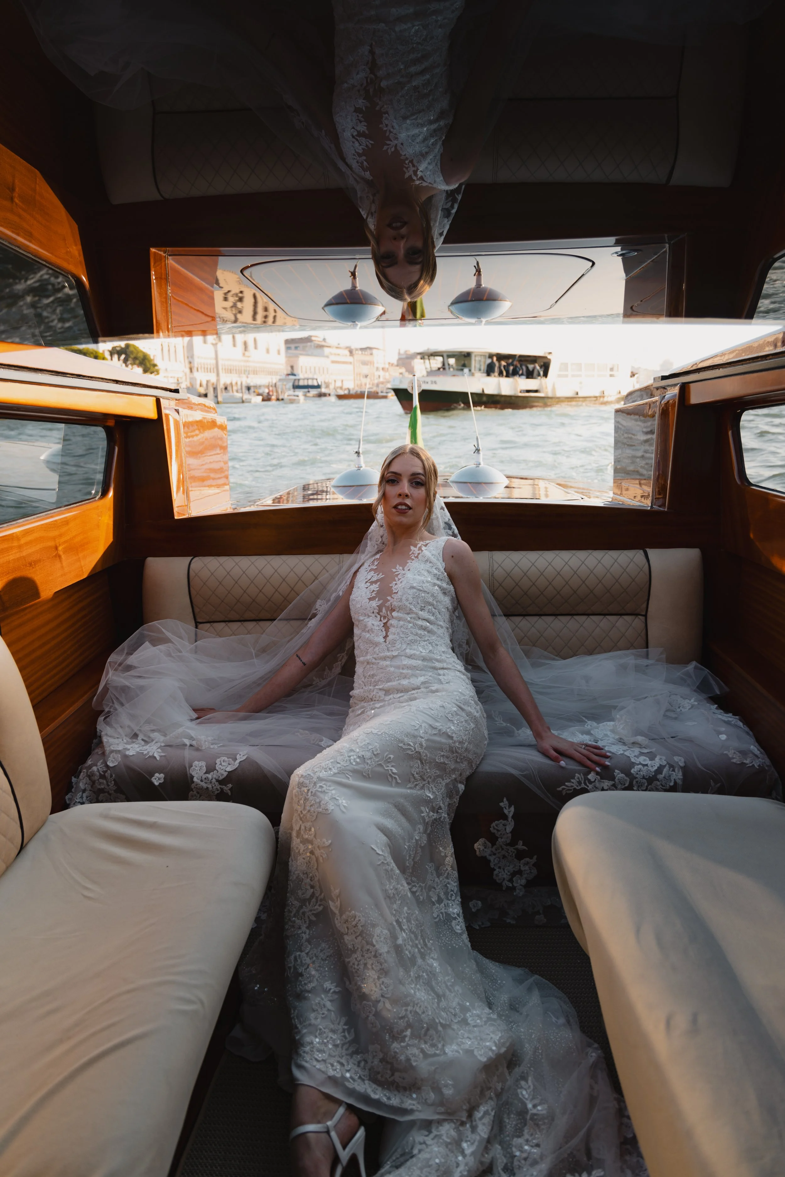 A bride in a white lace wedding dress sitting inside a boat with wooden paneling, nearby water, and other boats and buildings in the background.