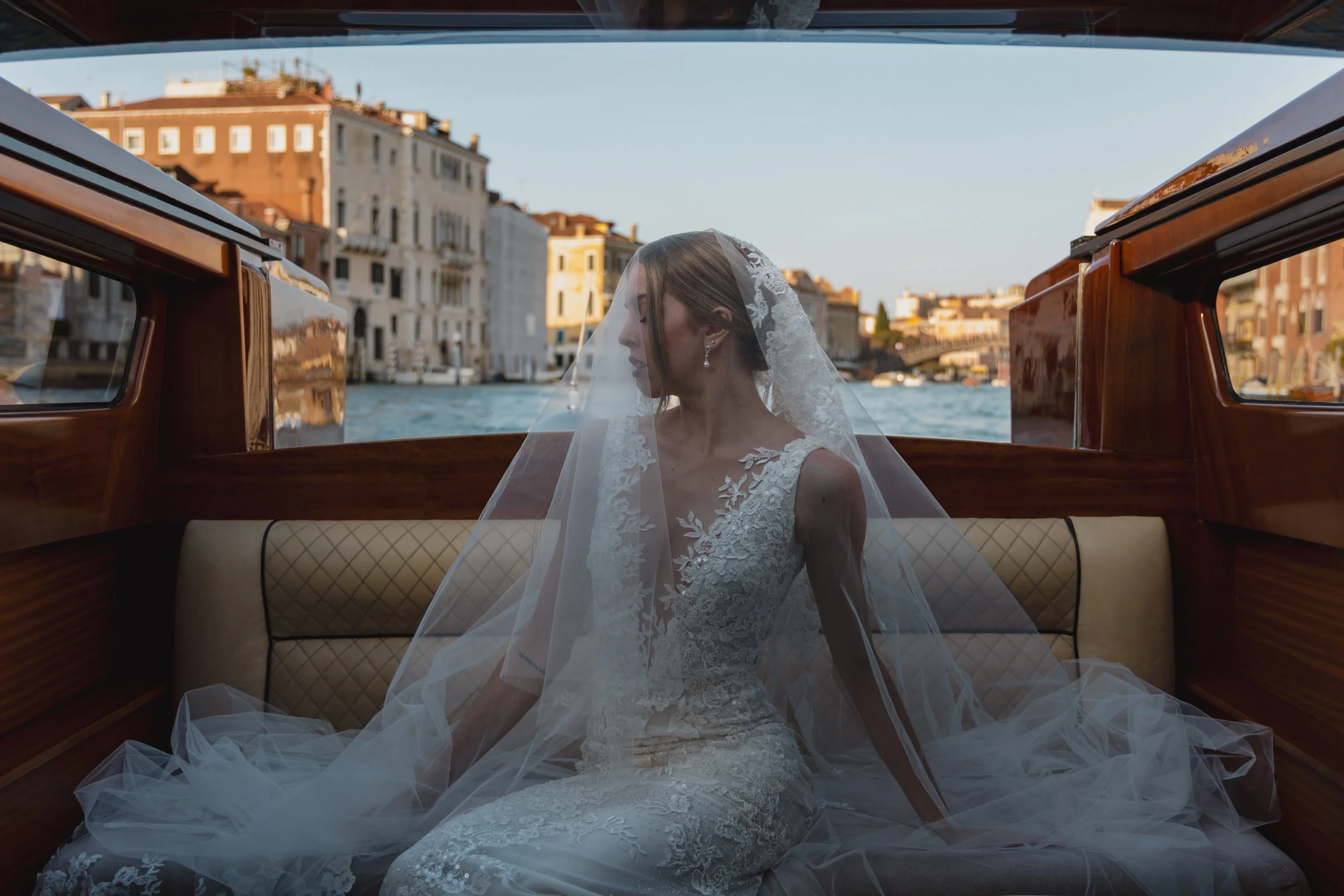 A bride in a white lace wedding gown and veil sitting inside a boat on a canal, with historic buildings in the background.