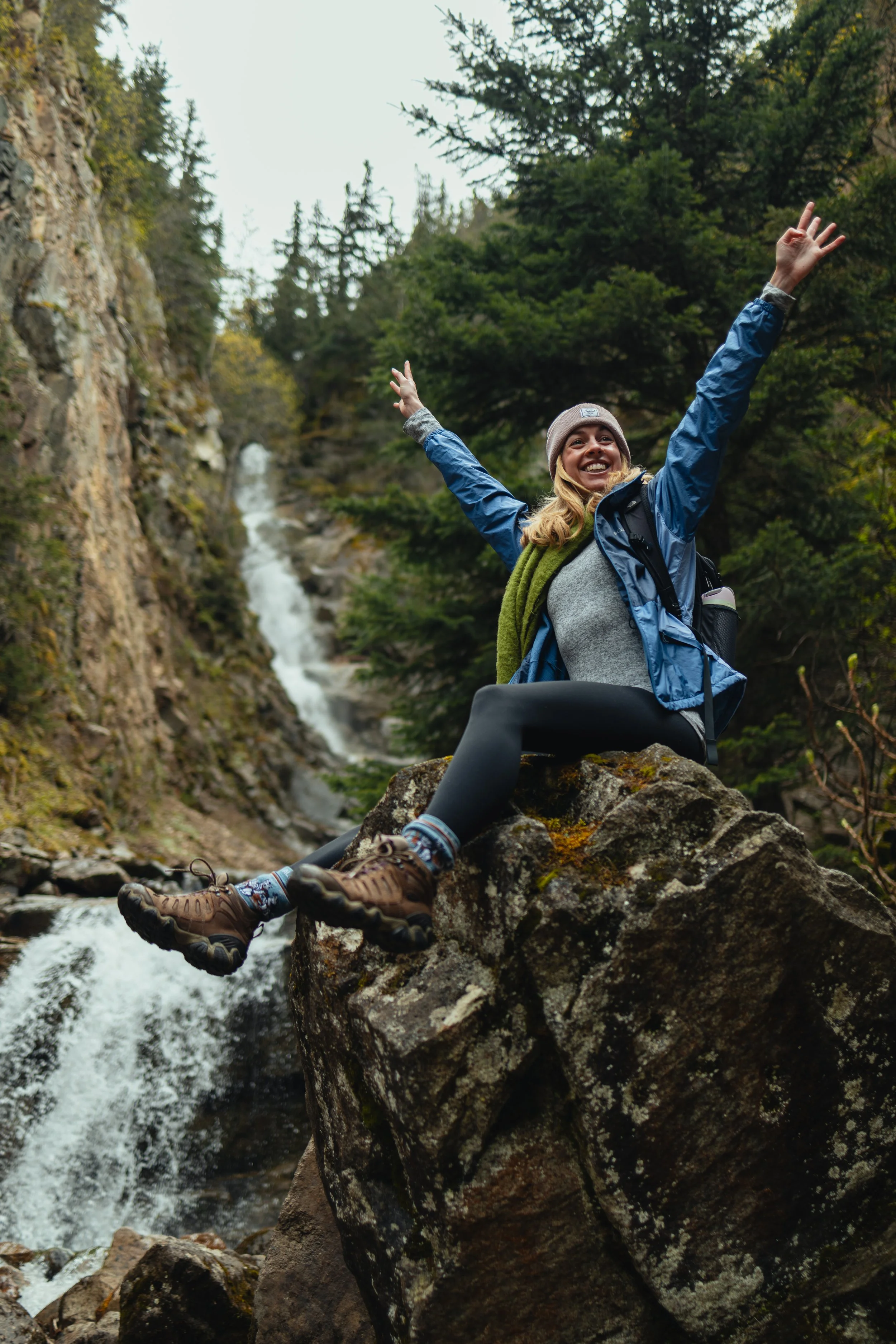 A woman smiling and joyfully raising her arms while sitting on a large rock by a waterfall surrounded by trees in a forest, wearing outdoor hiking gear.