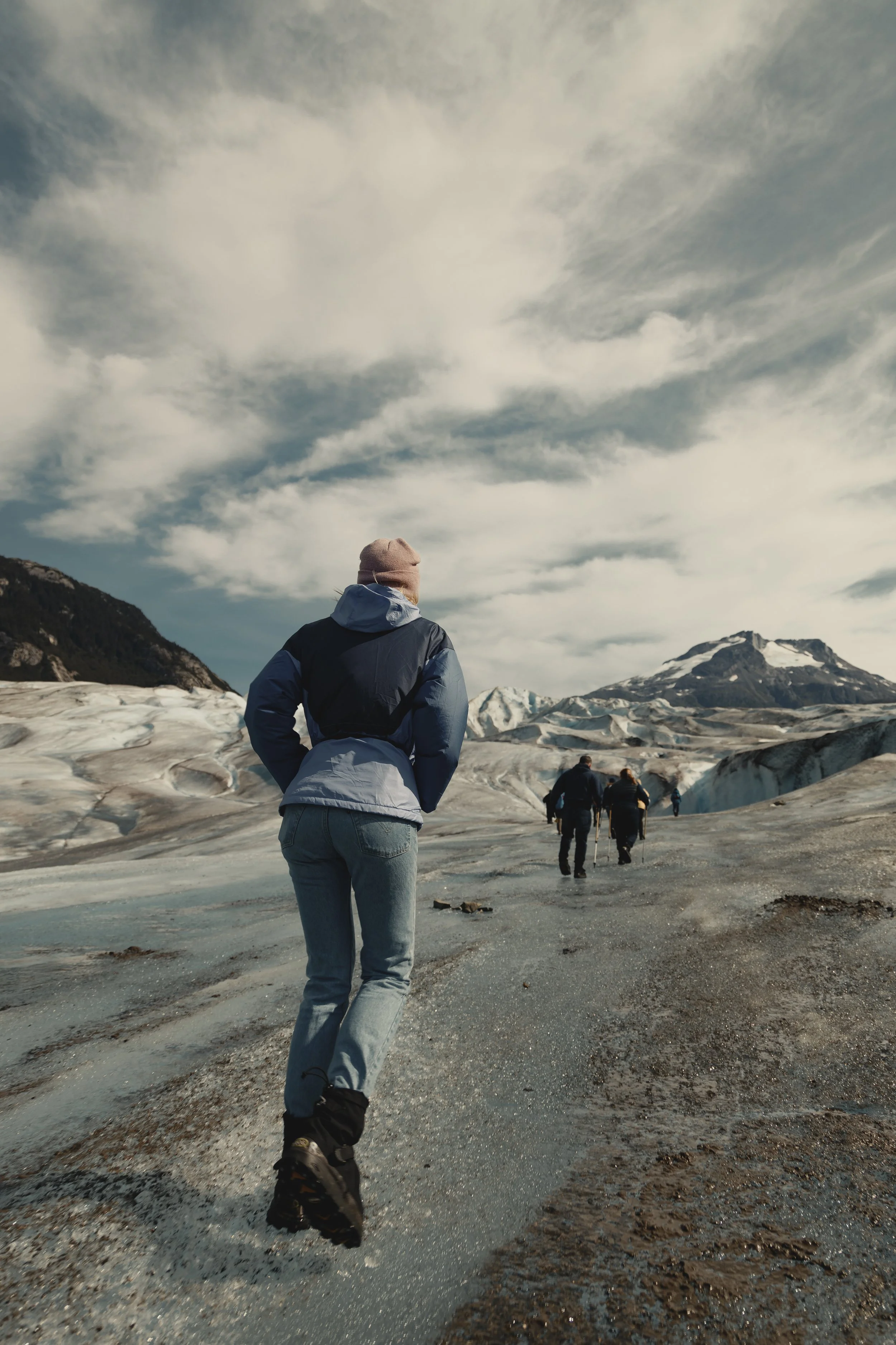 A person in a blue jacket and gray pants walking across a glacier with snow-capped mountains in the background.
