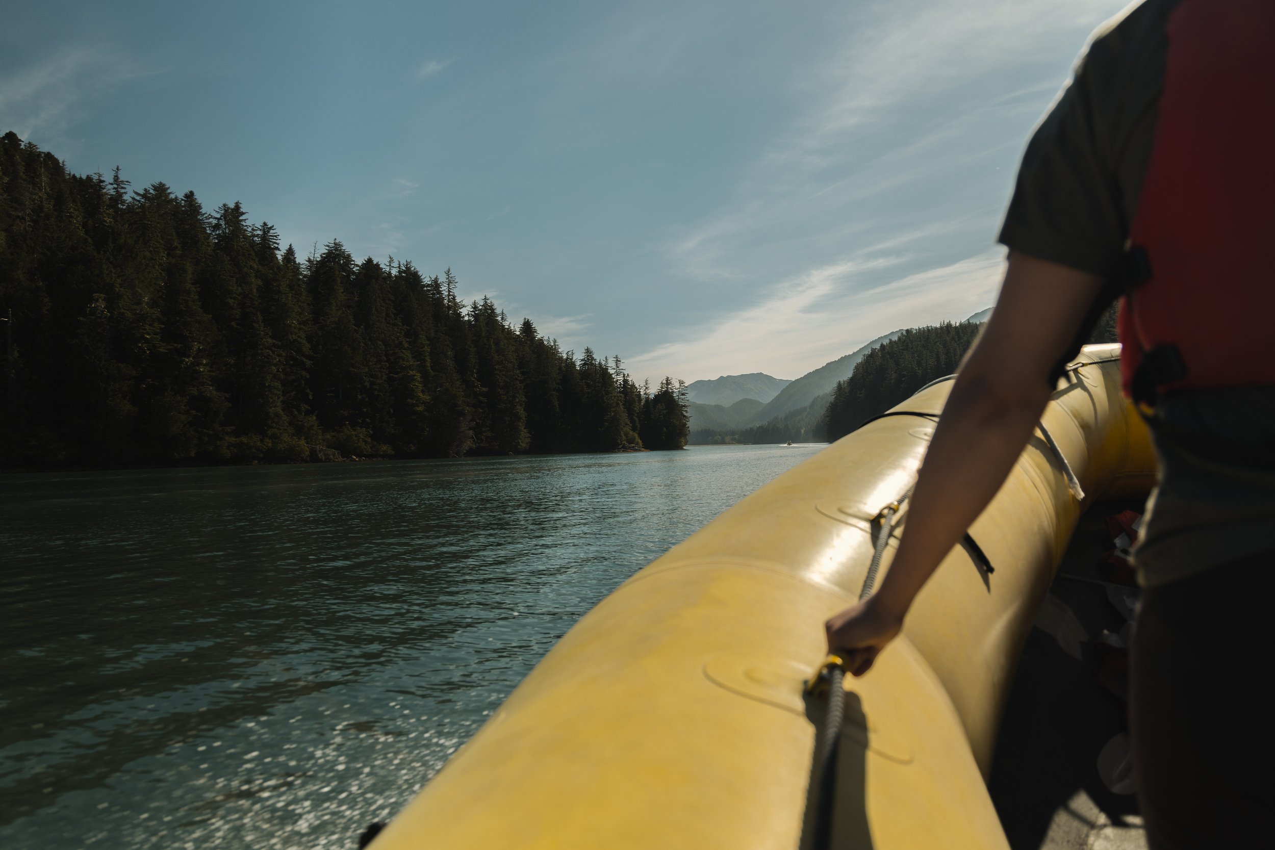 Person wearing a black and red life jacket paddles a yellow kayak on a river surrounded by tall green trees and mountains under a partly cloudy sky.