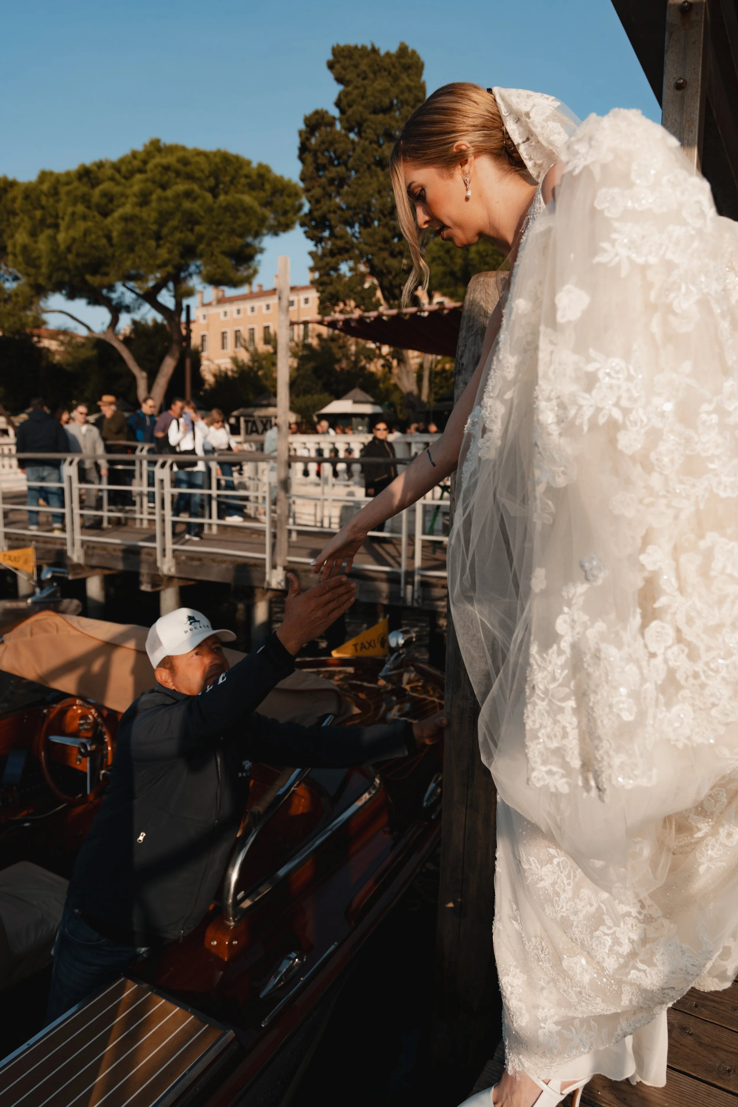 A bride on a dock wearing a white lace wedding dress, reaching out to a man in a black jacket and white cap sitting in a boat, with a group of people and trees in the background during daytime.