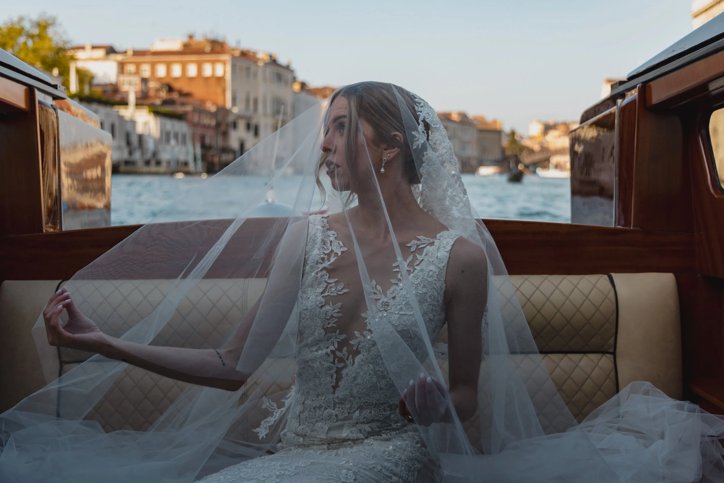 A bride in a lace wedding dress and veil sitting on a boat in a canal, with multi-story buildings in the background under a clear sky.